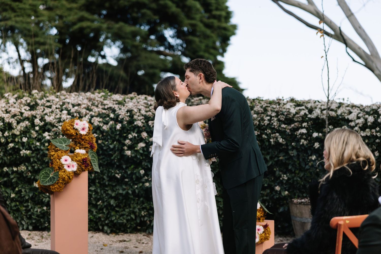 A bride and groom share their first kiss at an outdoor wedding ceremony with a lush floral hedge backdrop.
