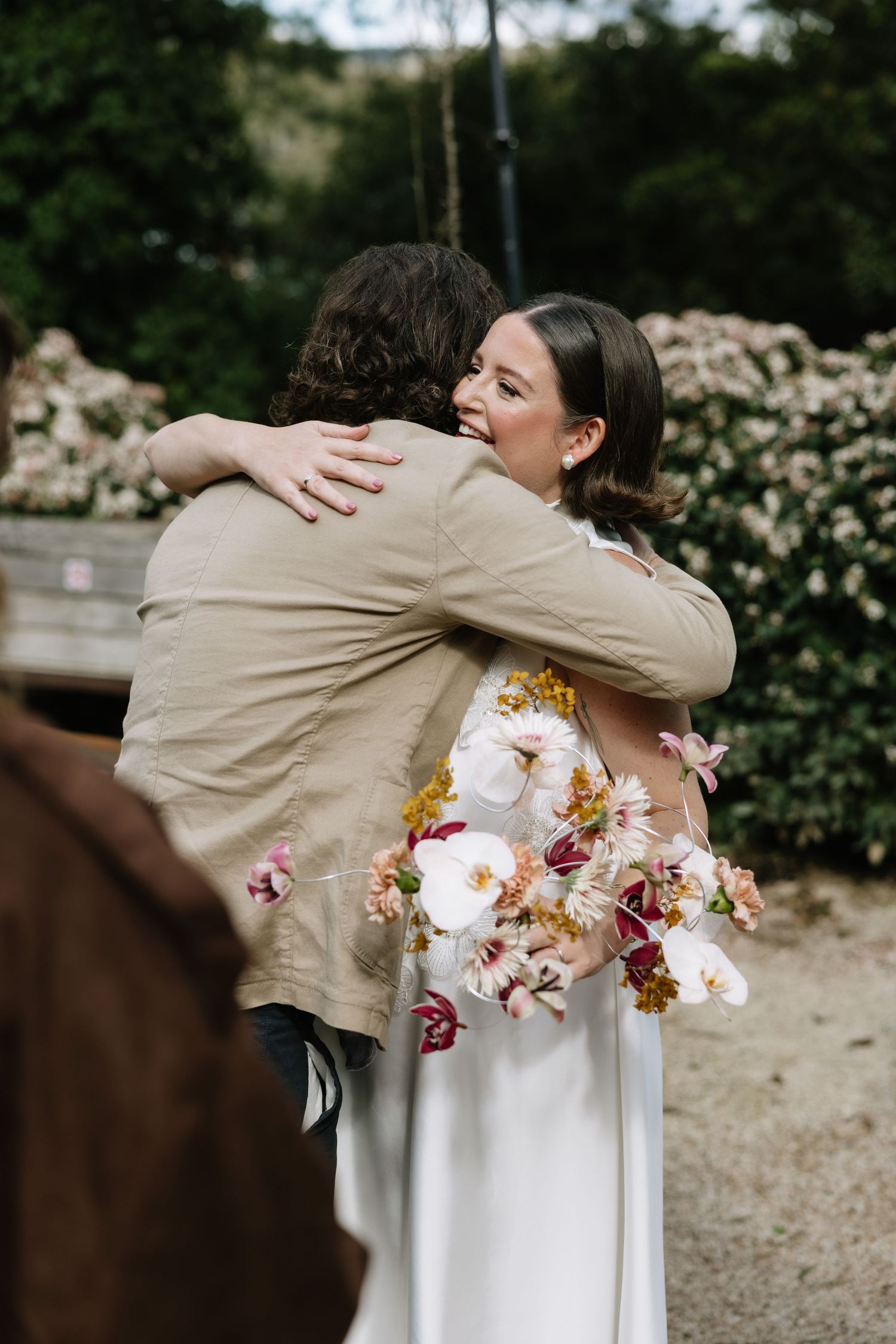 Bride in floral dress hugs guest while holding colorful bouquet outdoors at wedding venue.