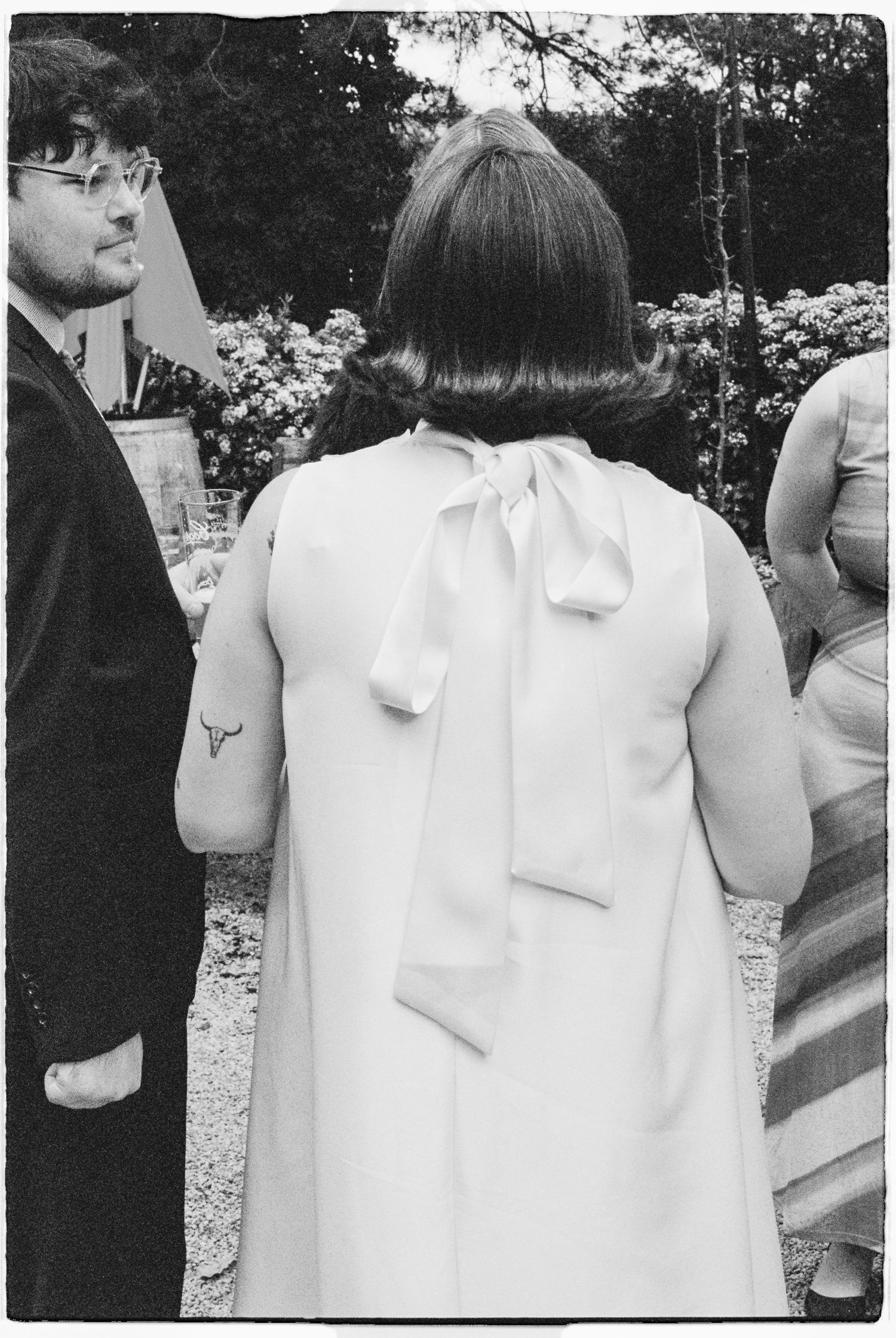 Black and white photo of a woman in a white bow-back dress standing outdoors at a garden event.