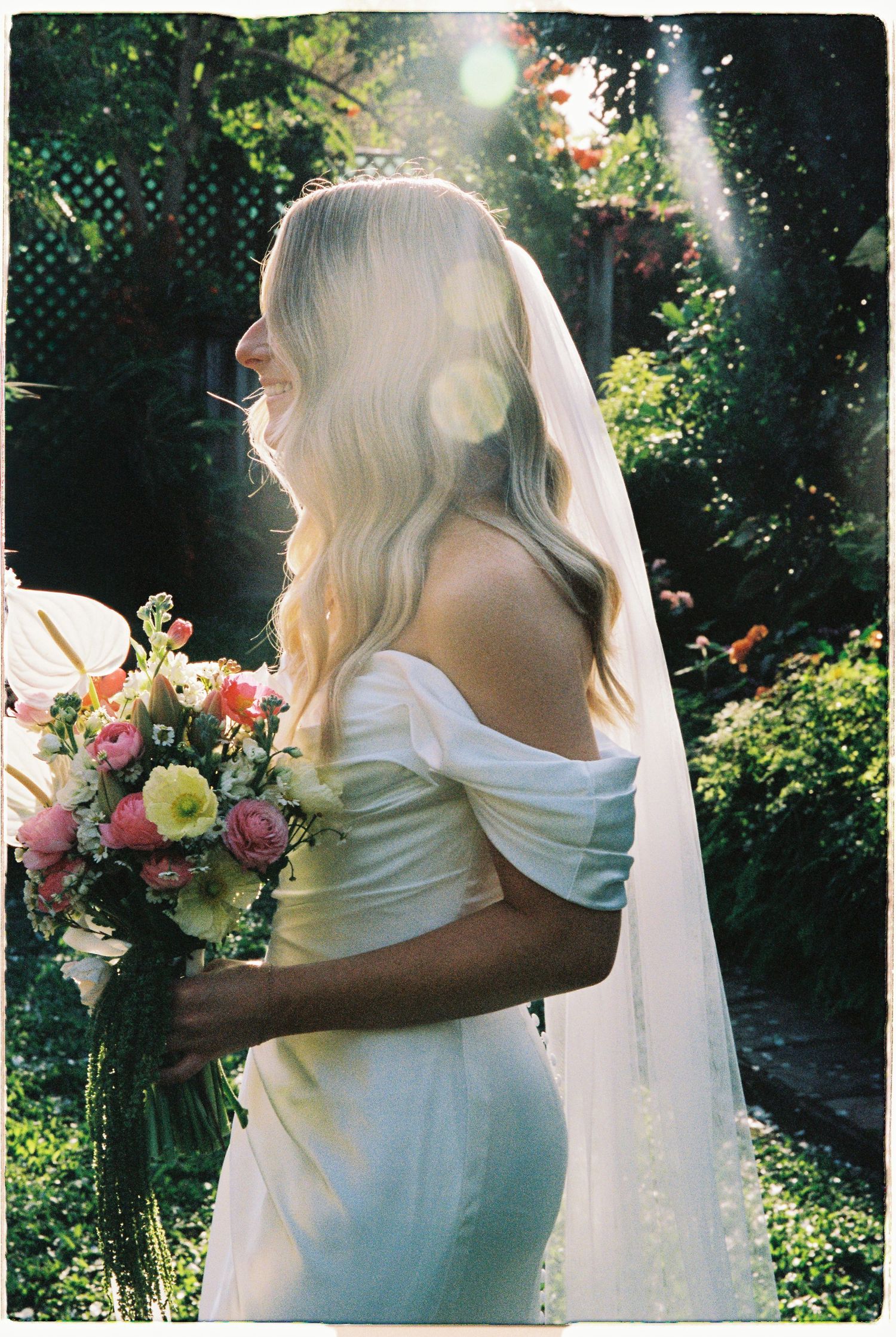 Bride in off-shoulder white gown and veil holds colorful bouquet in sunlit garden.