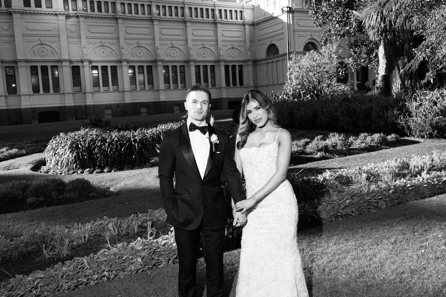 Black and white wedding photo of bride and groom holding hands outdoors in front of an elegant building.
