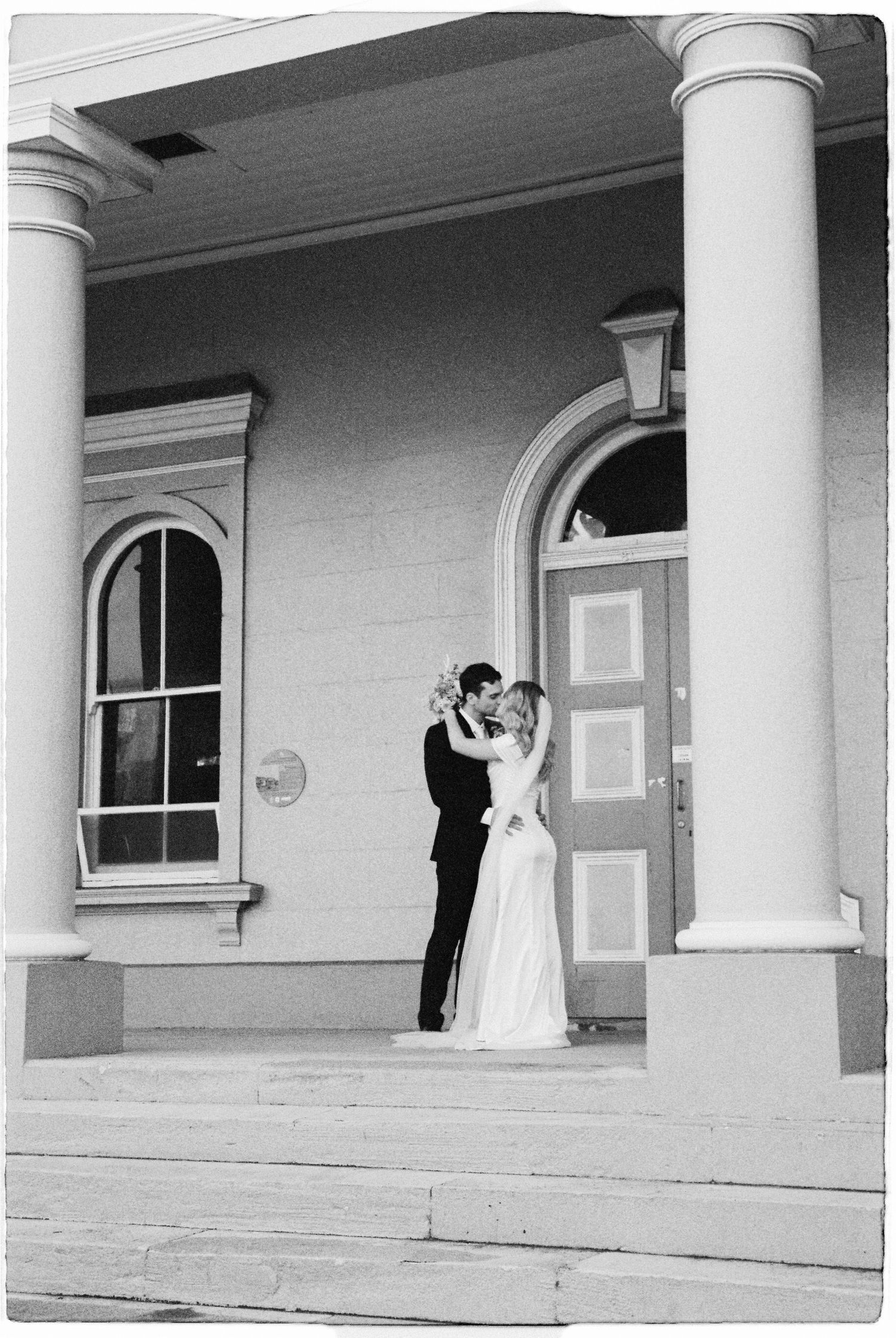 Black and white photo of bride and groom sharing a kiss on steps of historic building.