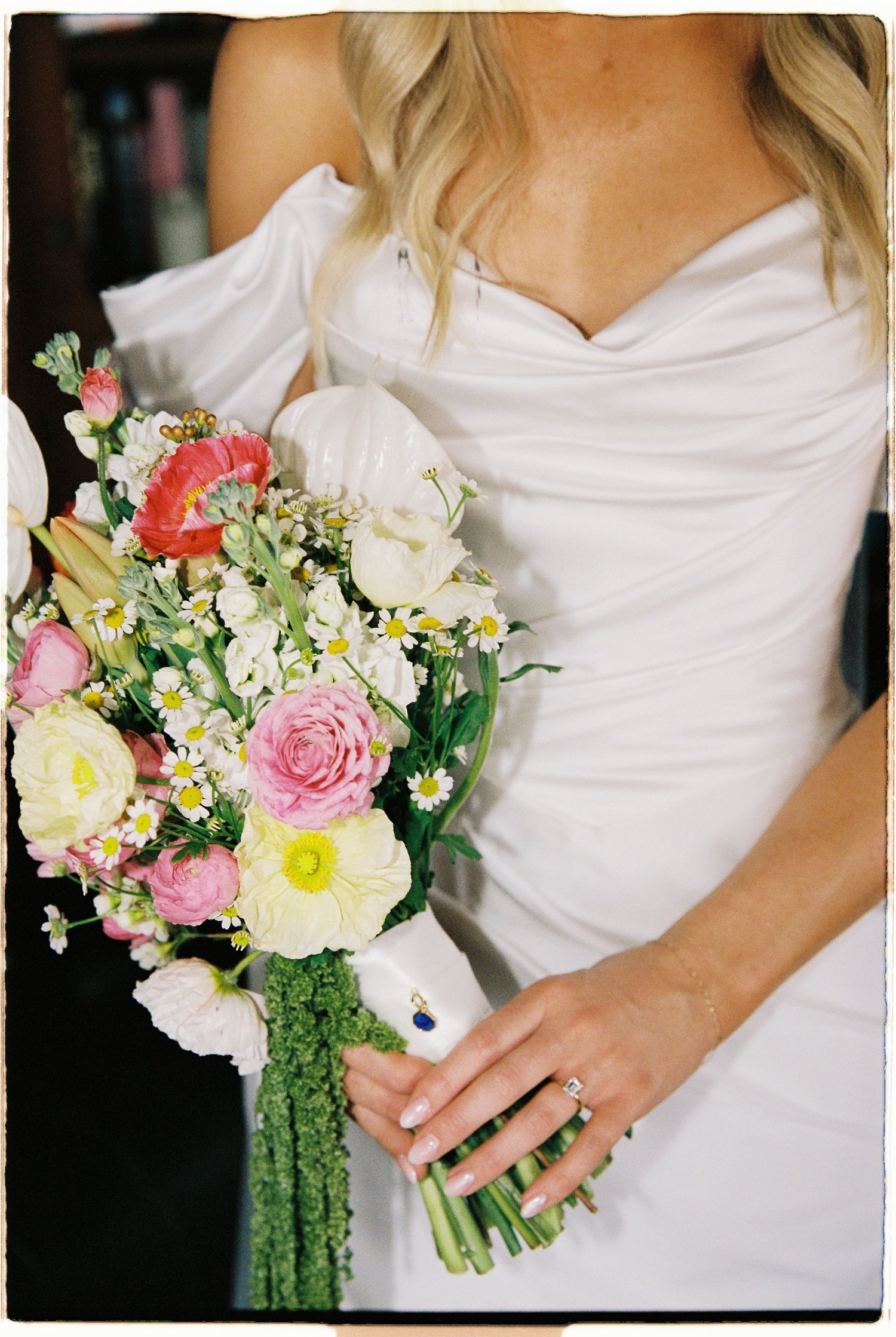 Bride in white off-shoulder gown holding a colorful bouquet of pink roses, daisies, and wildflowers.