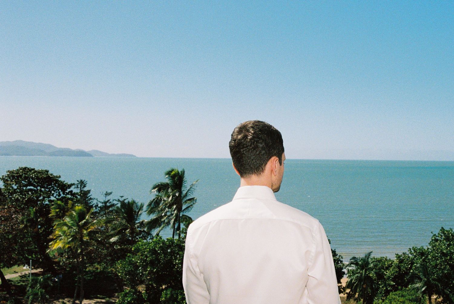 Man in white shirt gazing at tropical ocean view with lush green palm trees under clear blue sky.