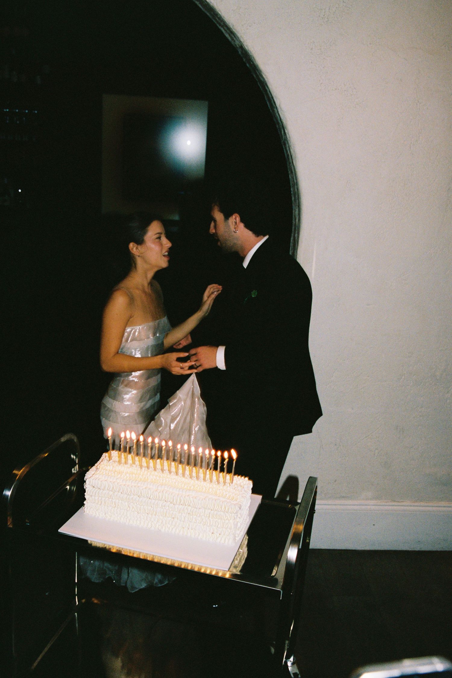 Couple cutting a white wedding cake with lit candles in elegant formal attire at a reception.