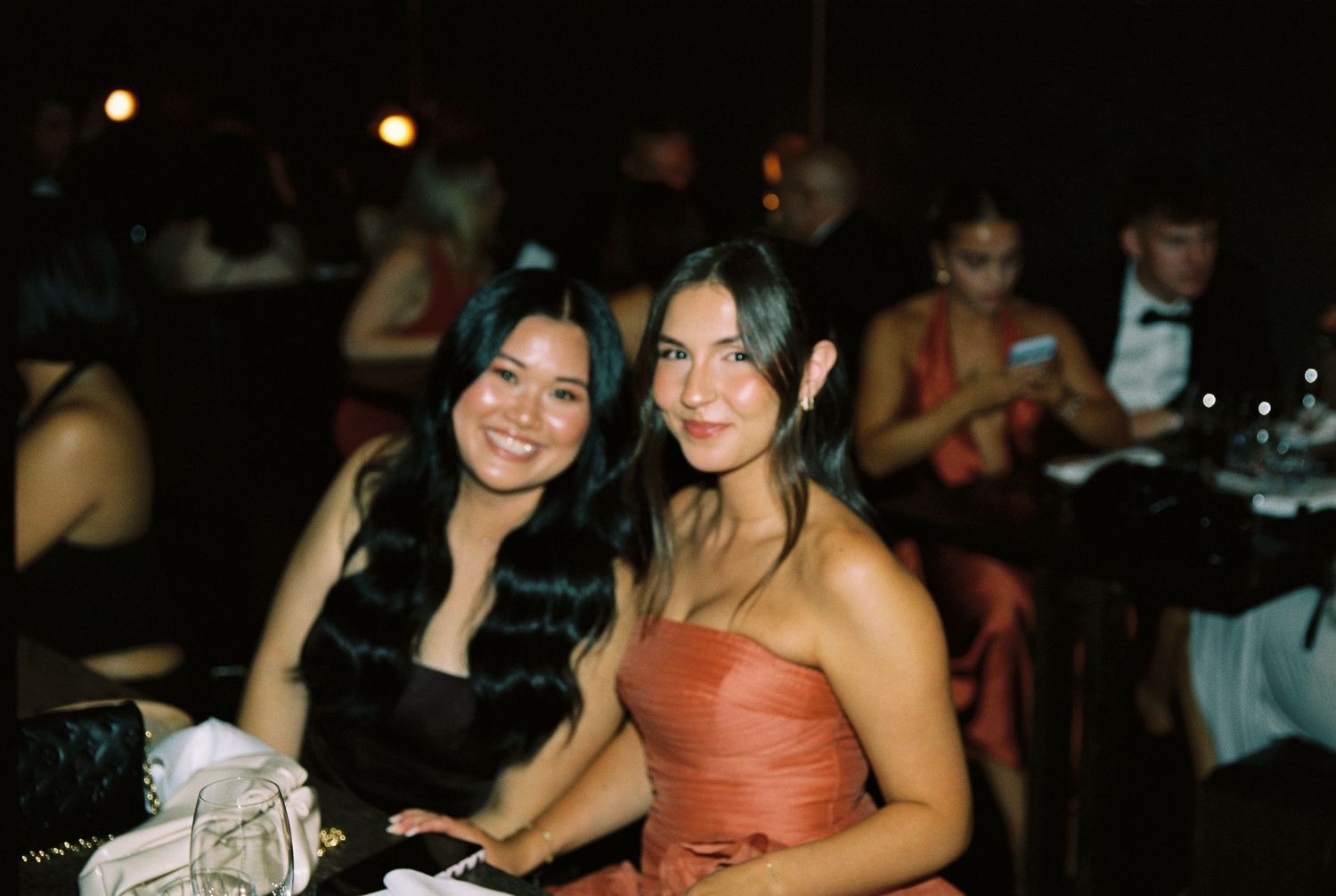 Two women in formal attire smile together at a dark, elegant wedding reception venue.