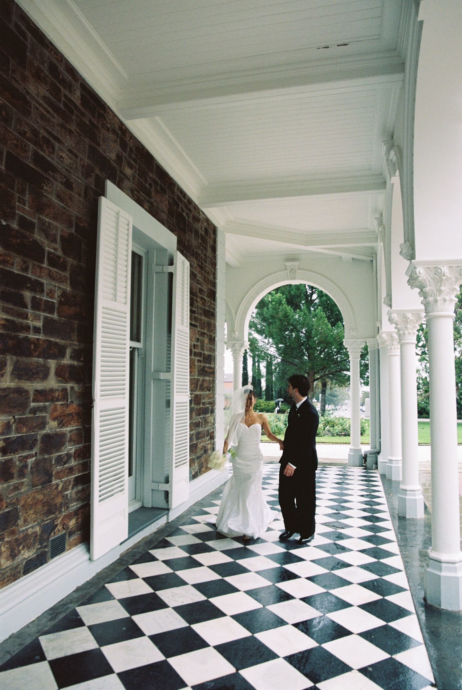 Bride and groom share a romantic moment on a elegant checkered porch with white columns at a historic venue.