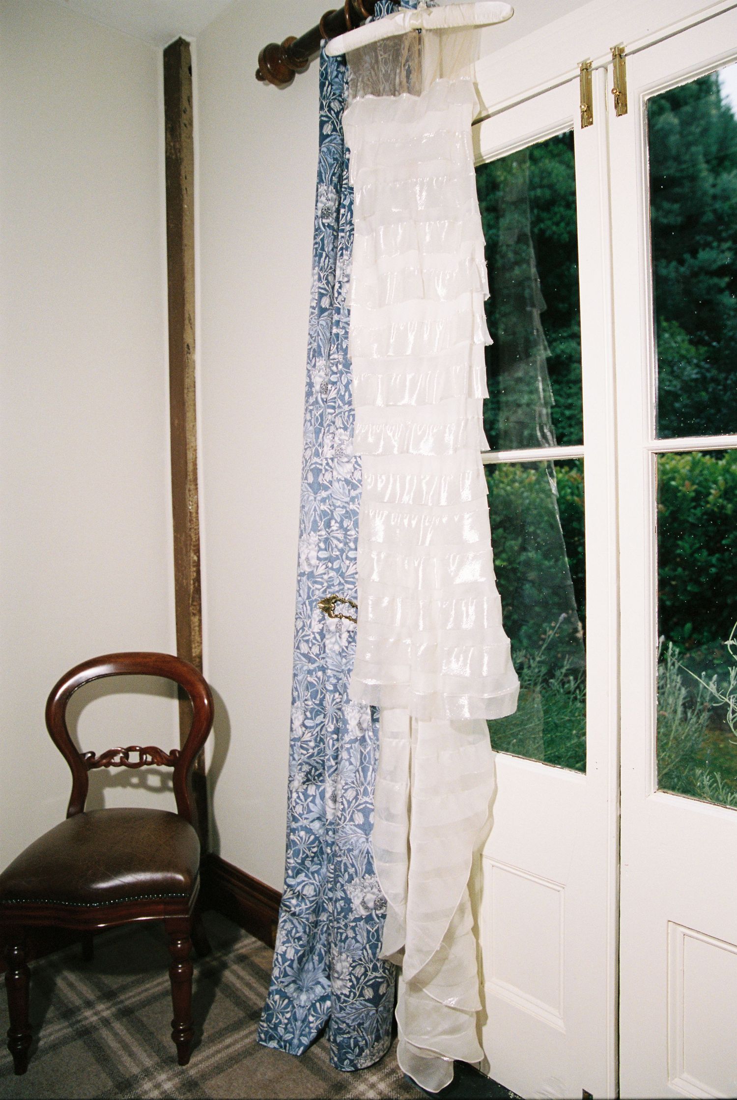 Wedding dress and blue floral sash hanging on curtain rod beside white door with garden view.
