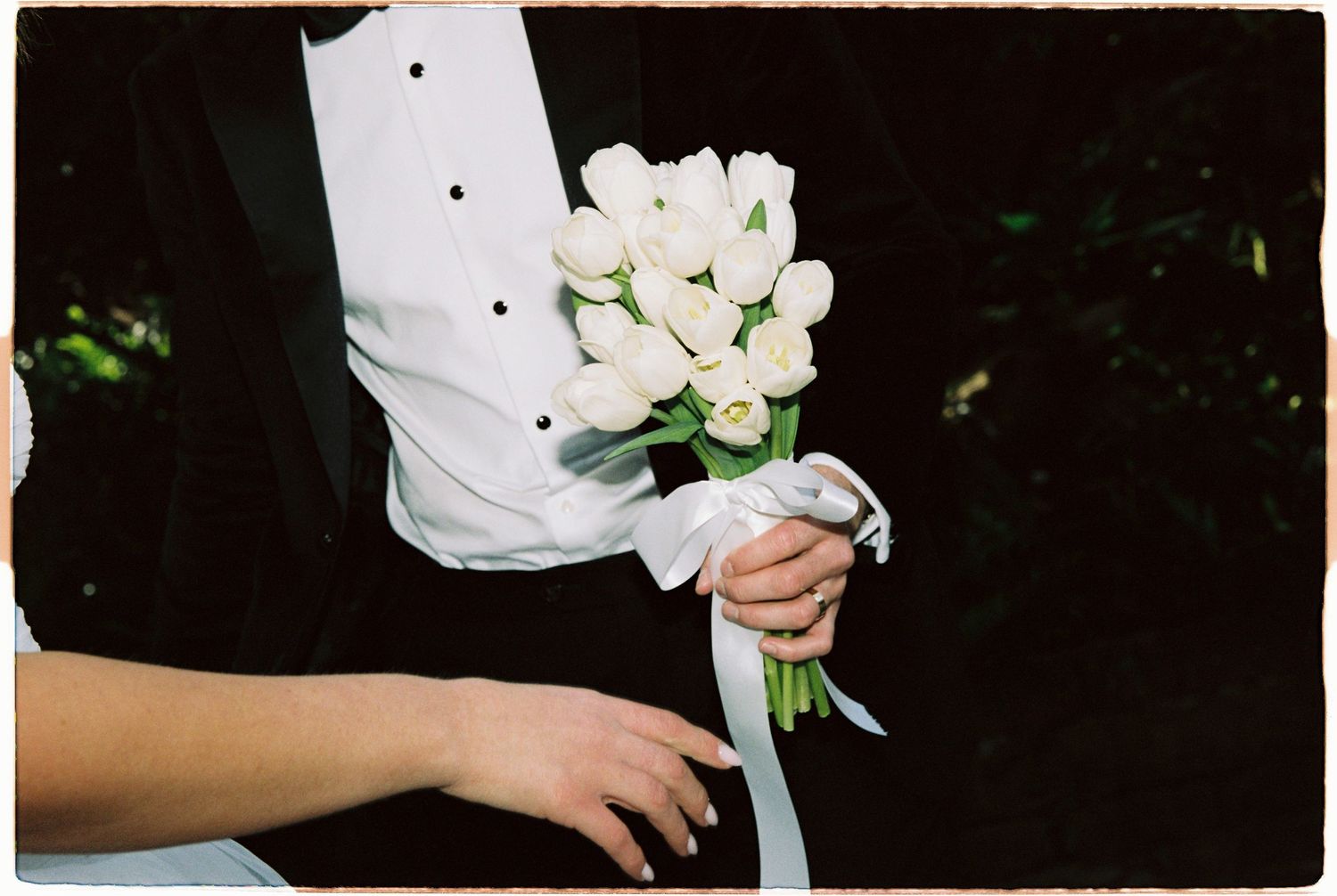 Groom in black tuxedo holding a white tulip bridal bouquet tied with ribbon.