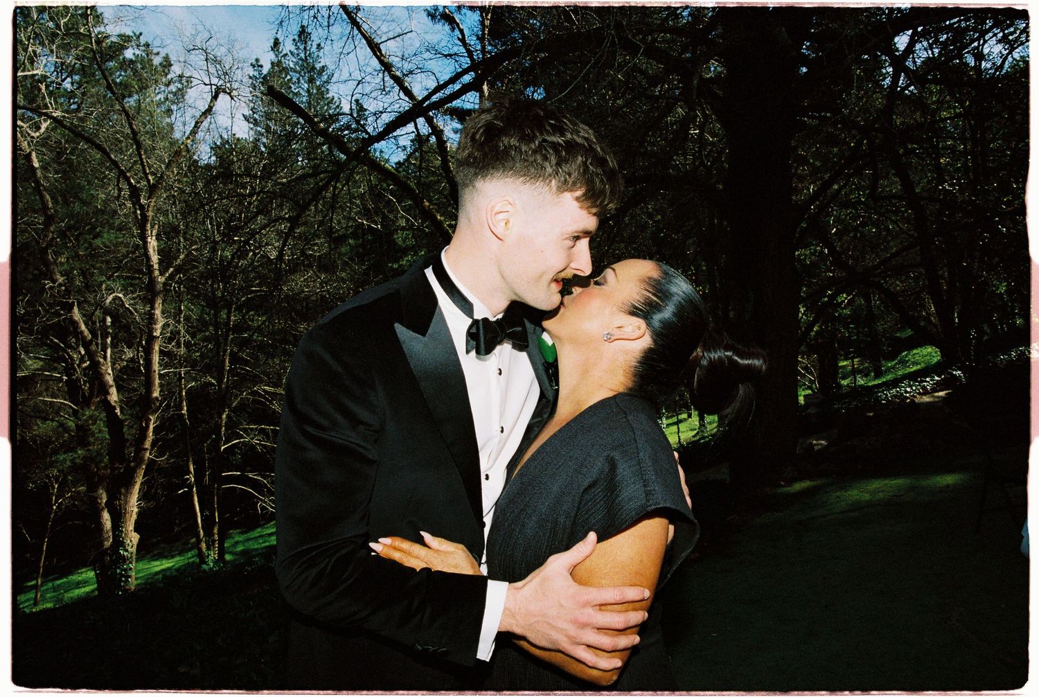 Couple in formal attire sharing a romantic kiss surrounded by lush green trees.