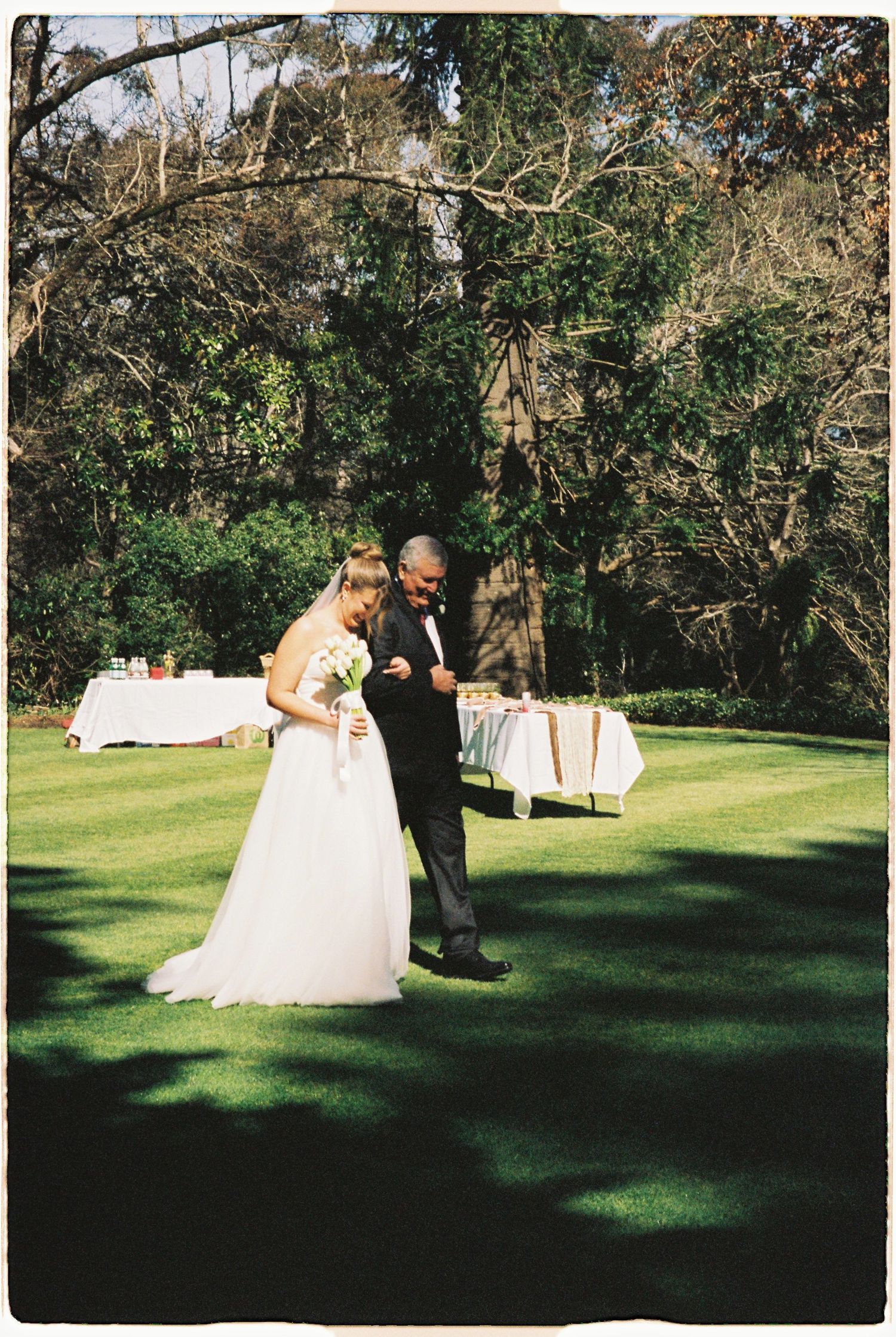 Bride in white gown walks with groom in black tuxedo across lush green lawn at outdoor wedding reception.