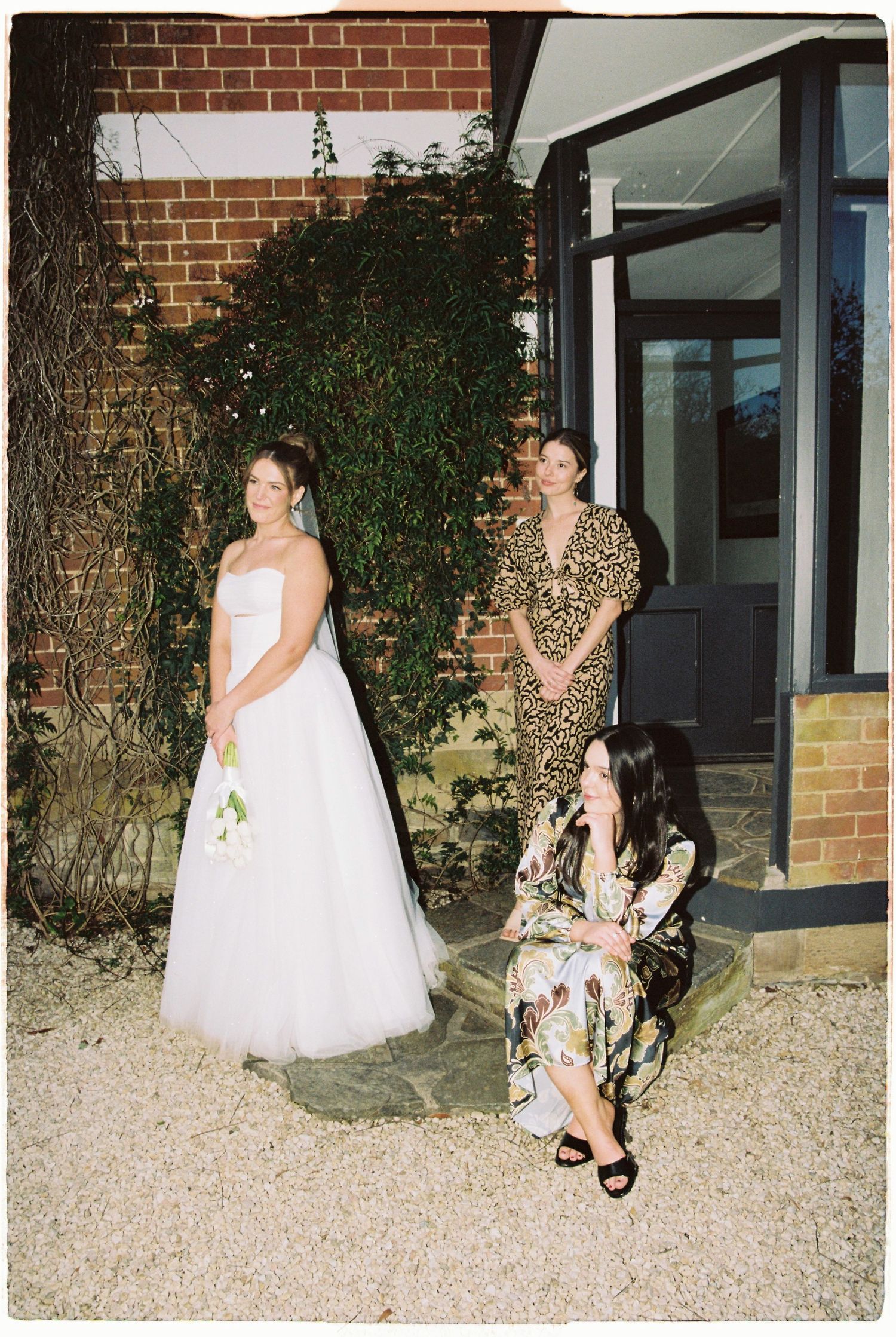 Bride in white strapless gown holds bouquet while two guests in floral dresses pose by brick wall.
