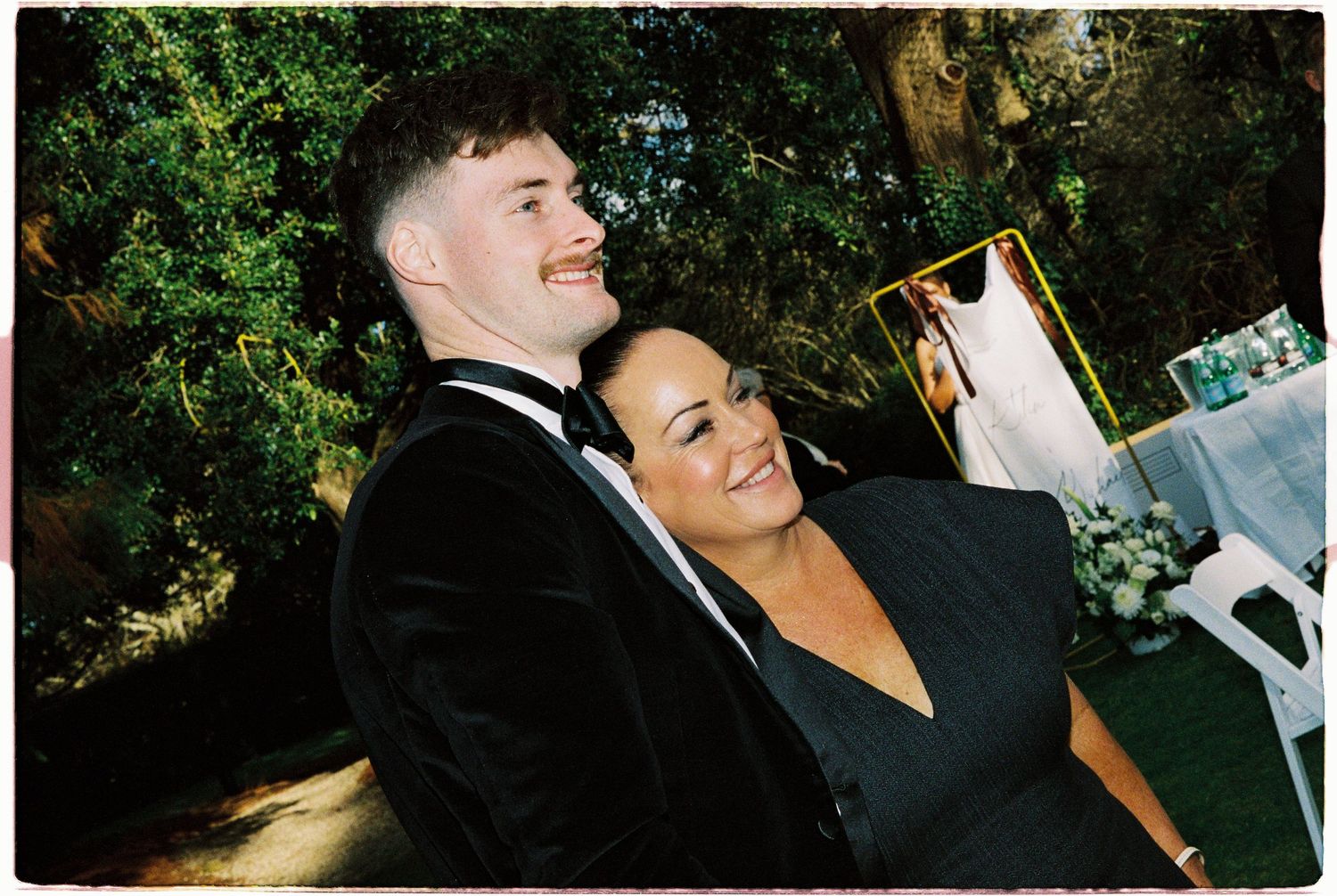 Groom in tuxedo and woman in black dress share a joyful moment together at an outdoor wedding reception.