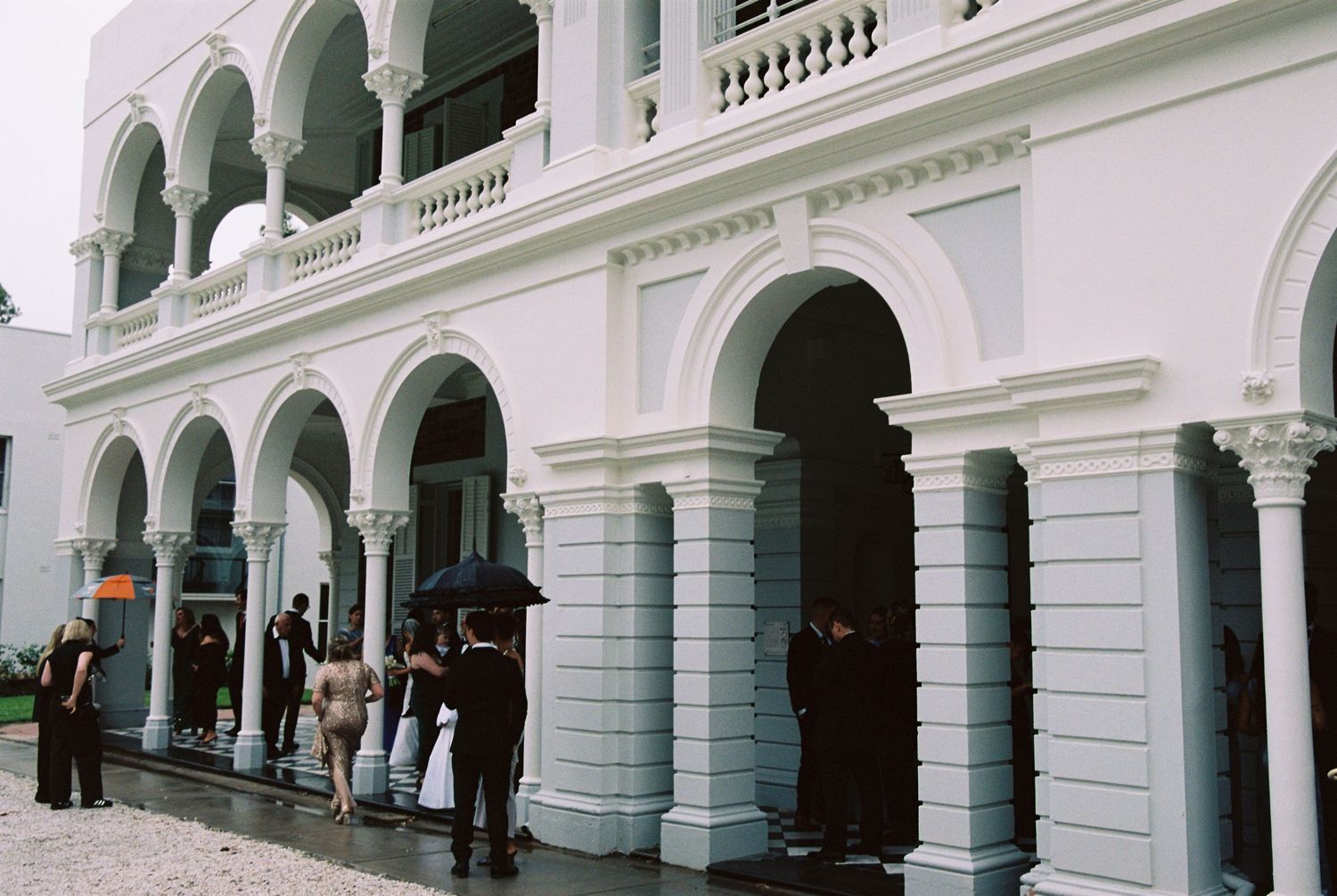 Wedding guests gather under arches of elegant white colonial building on rainy day.