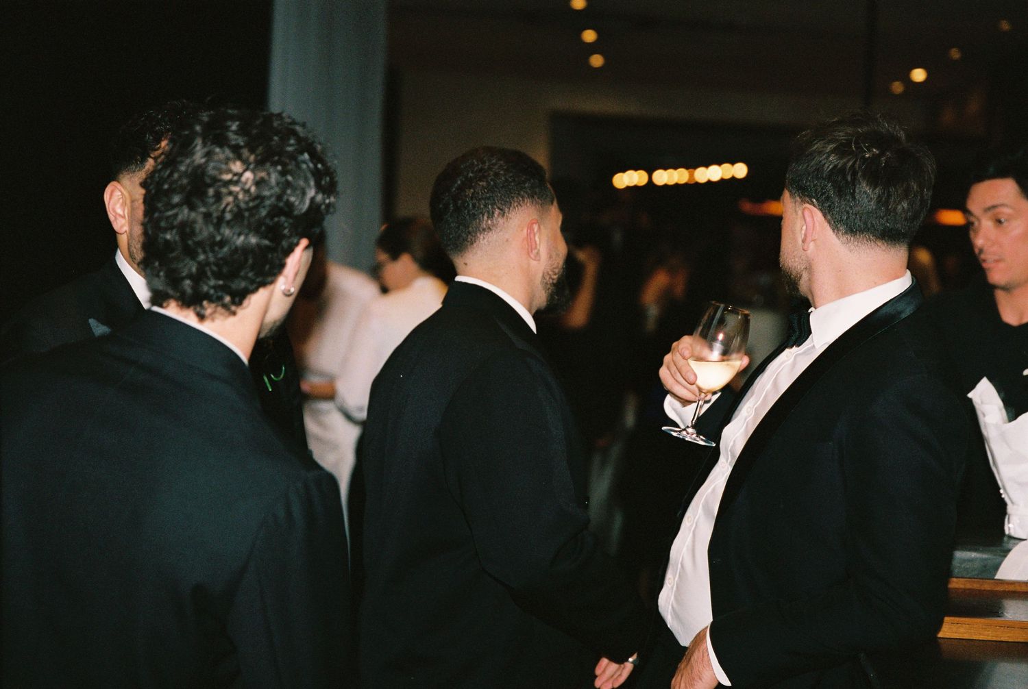 Three men in black tuxedos socializing at a wedding reception, one holding a champagne glass.