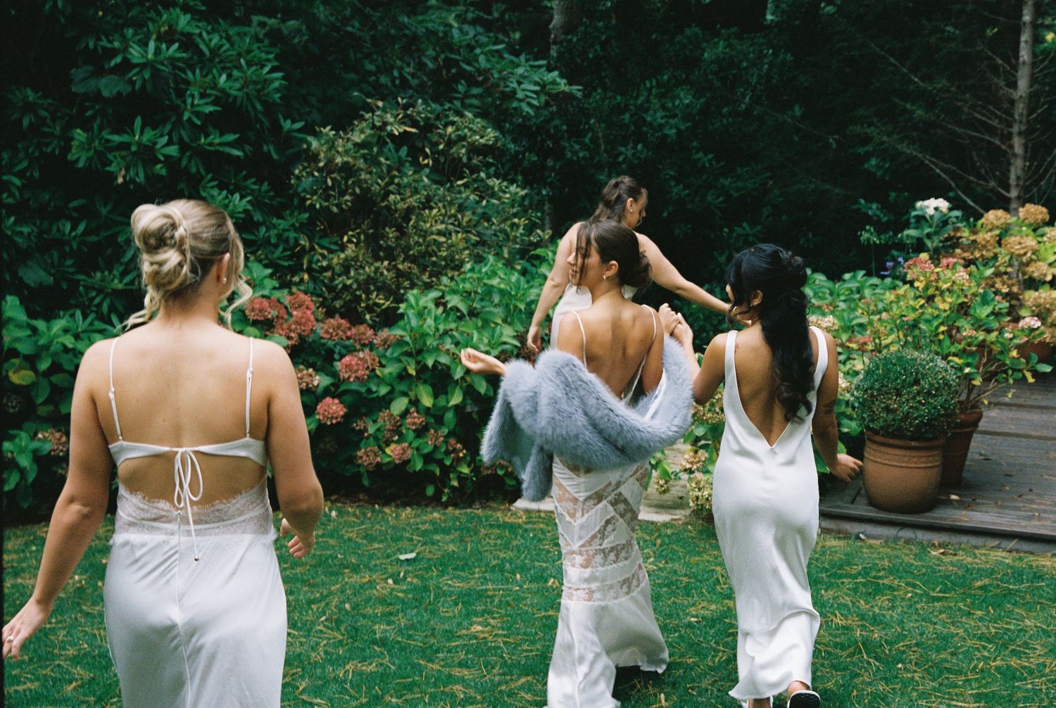 Four women in white dresses walk through a lush garden, captured from behind in a candid outdoor setting.