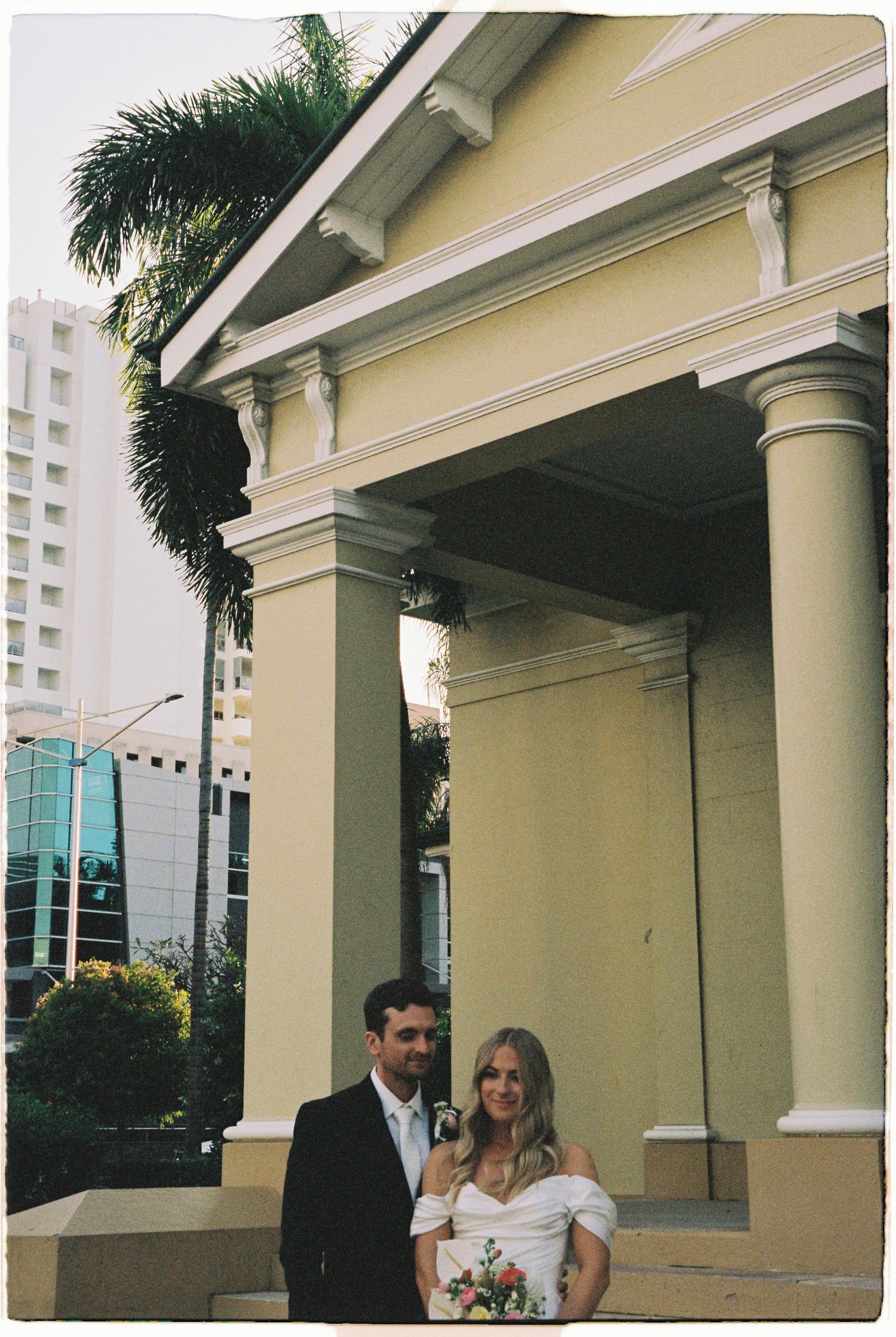 Bride and groom posing together in front of a classical colonial-style building with palm trees.