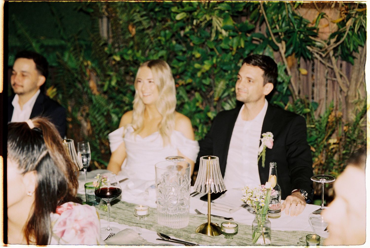 Bride and groom seated at elegant wedding reception dinner table with greenery backdrop and candlelight.