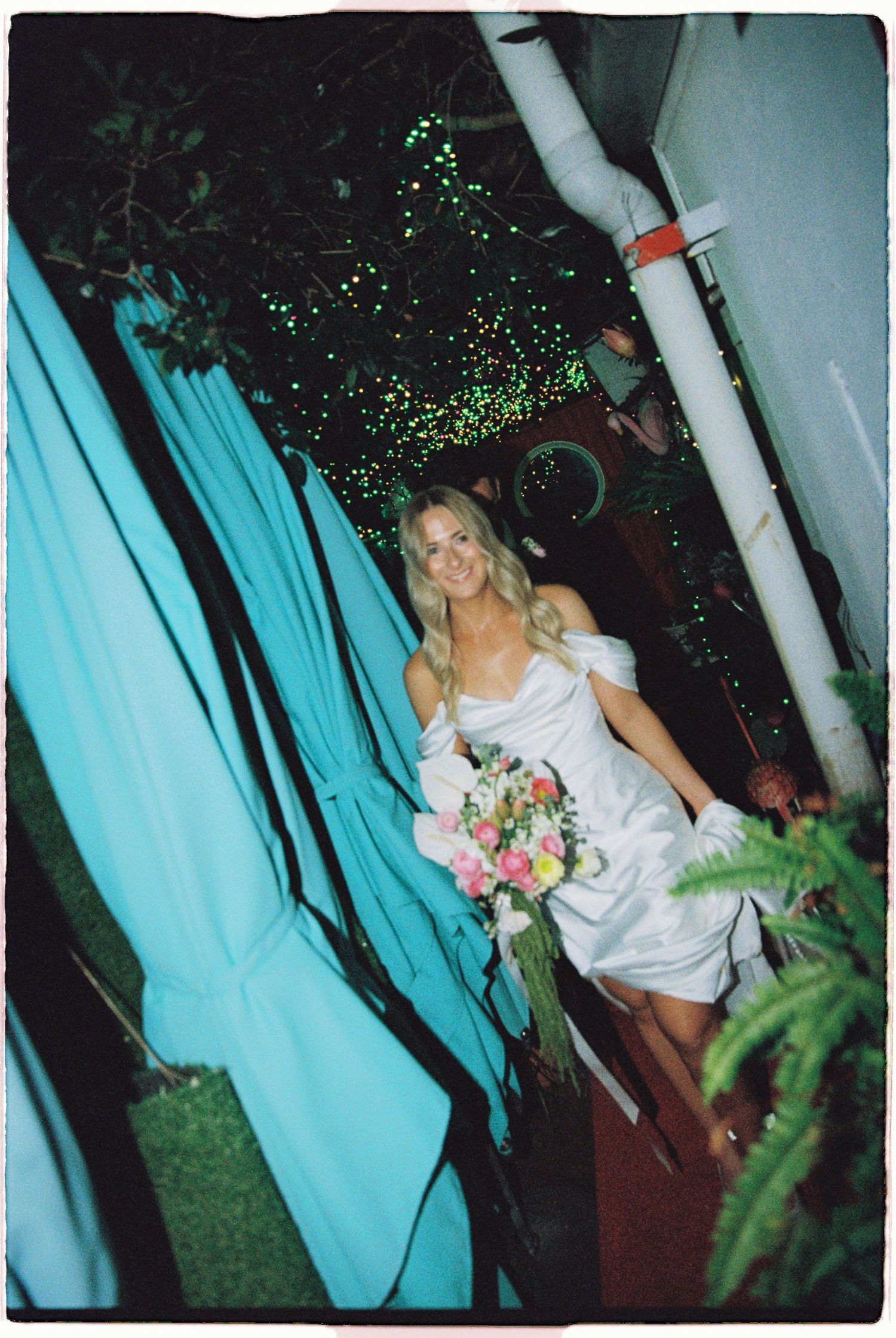 Radiant bride in white off-shoulder gown holding pink bouquet walking through teal draped entrance at night.