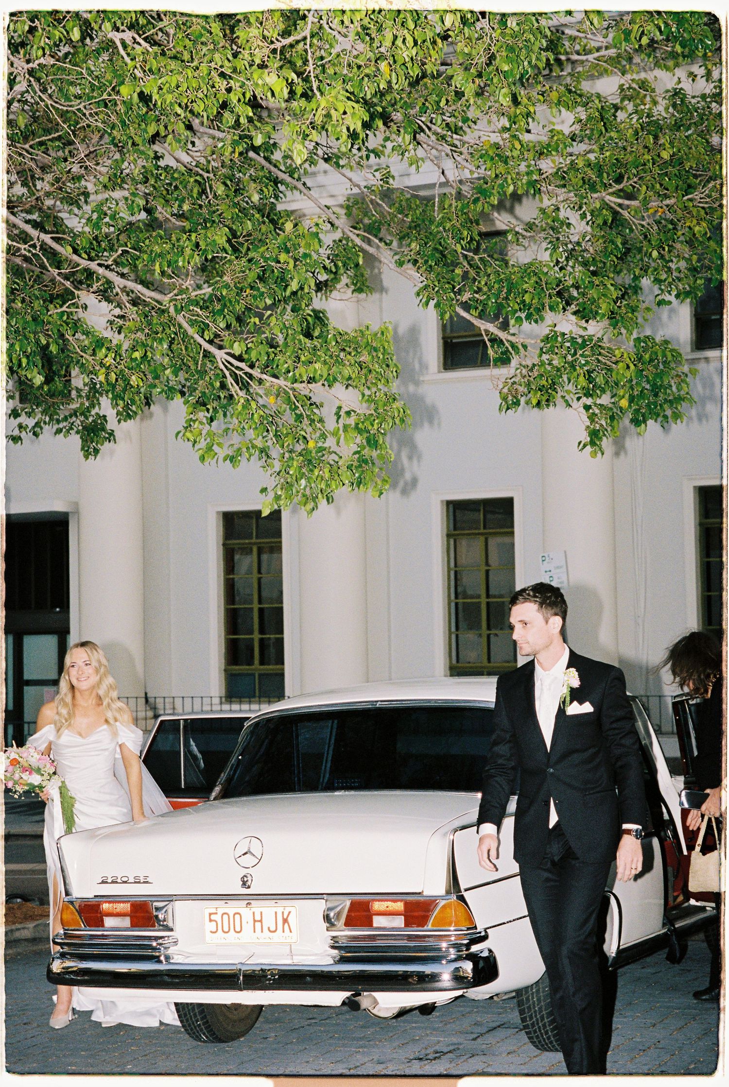 Bride and groom exit vintage white Mercedes-Benz 300SL outside elegant white classical wedding venue.