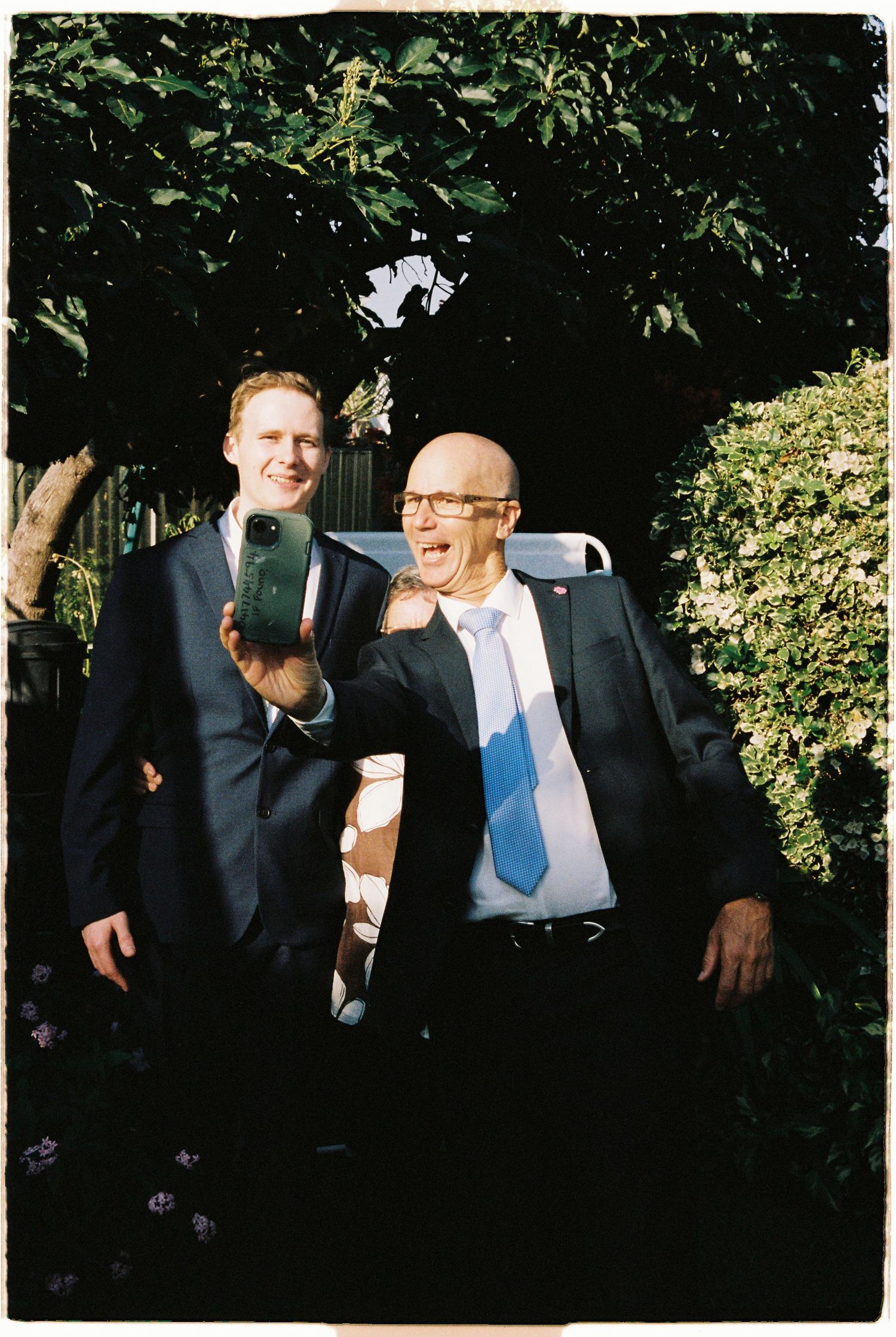 Two men in dark suits laugh together in a sunlit garden, one holding a smartphone.