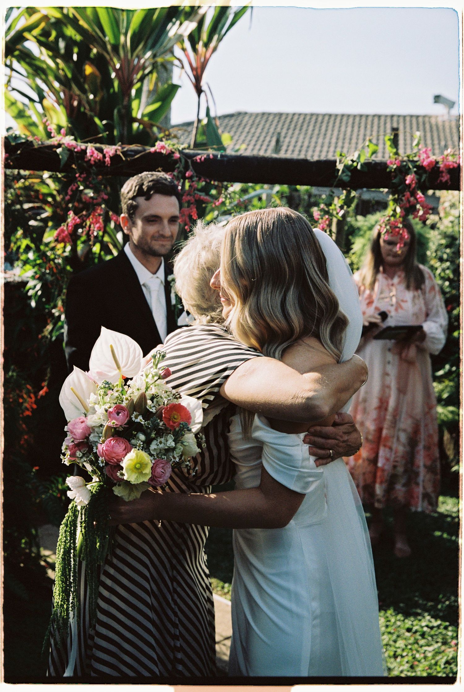 Bride hugging guest at outdoor garden wedding ceremony with groom smiling behind.