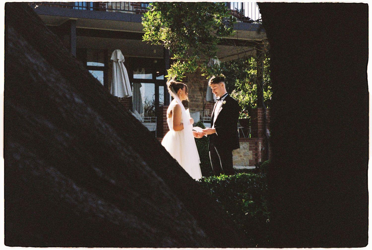 A bride and groom exchange vows in a sunlit outdoor ceremony beside a historic building surrounded by trees.
