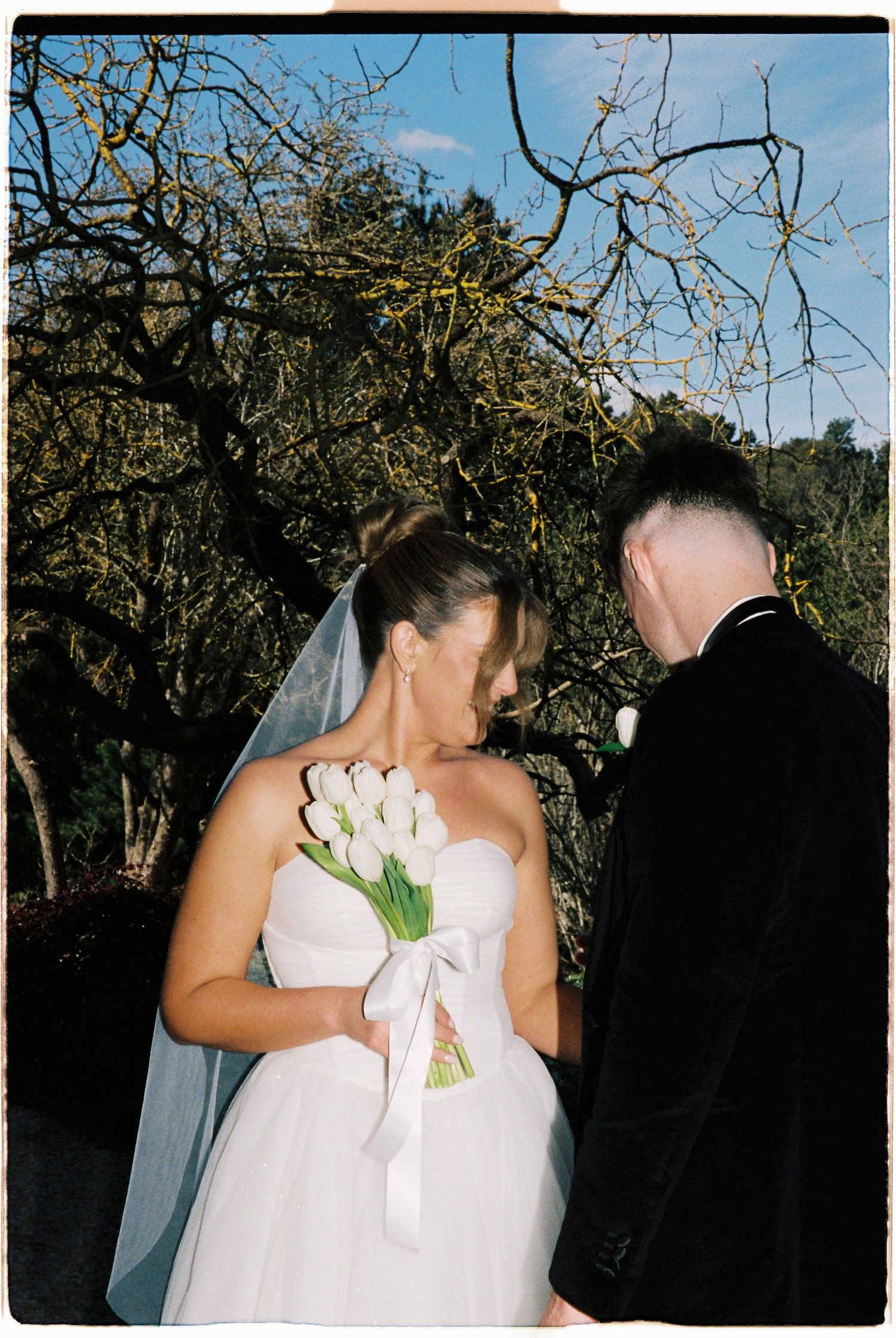 Bride in strapless white gown holding white tulip bouquet stands with groom outdoors.