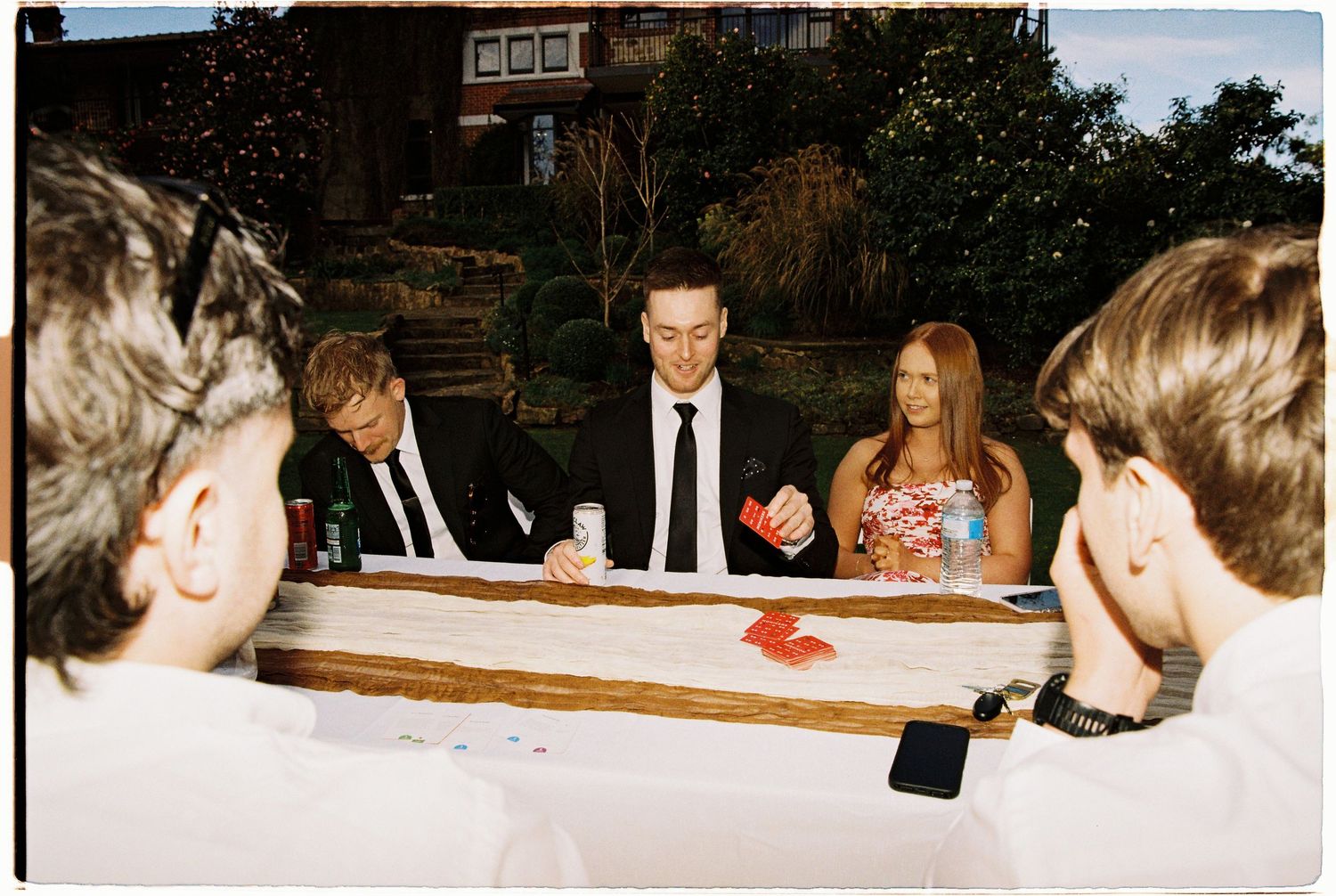 Wedding guests in formal wear gathered around a table playing a game at an outdoor reception.