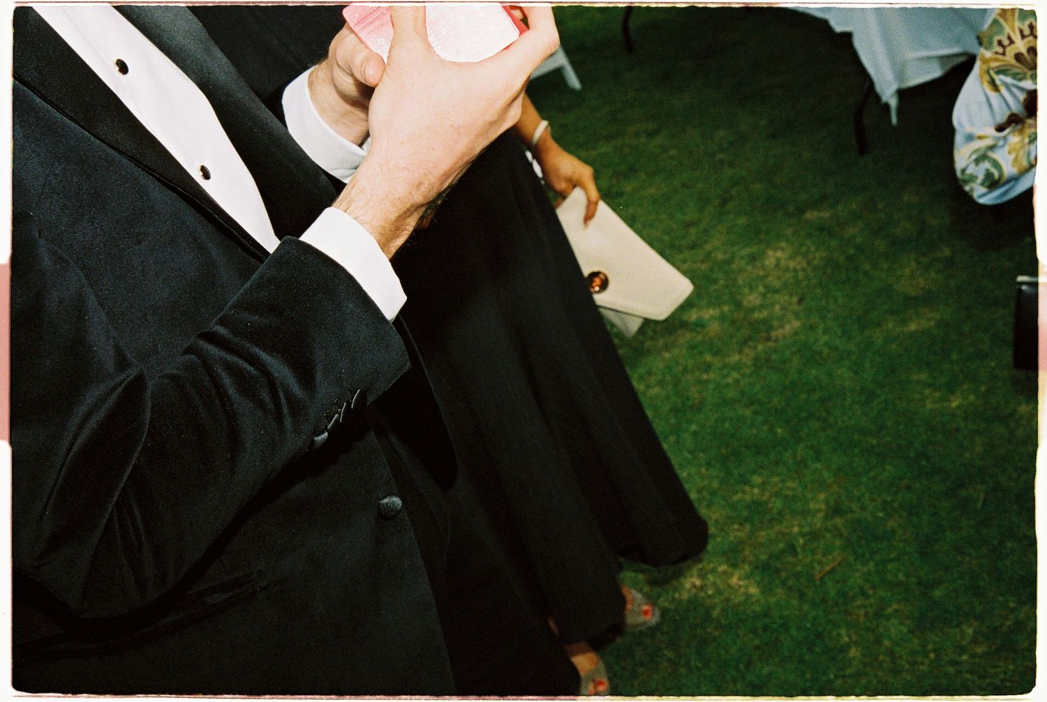 Close-up of wedding guest in black tuxedo holding a pink drink cup on a green lawn.