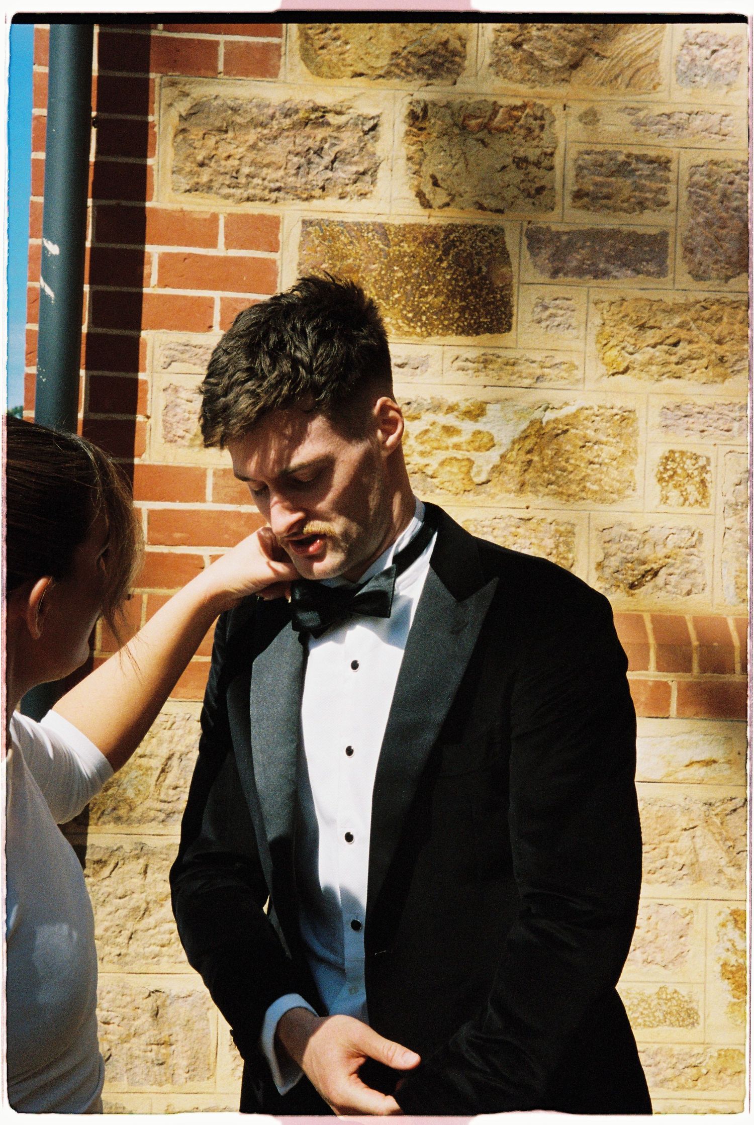 Groom in black tuxedo with bow tie standing against a stone wall while someone adjusts his collar.