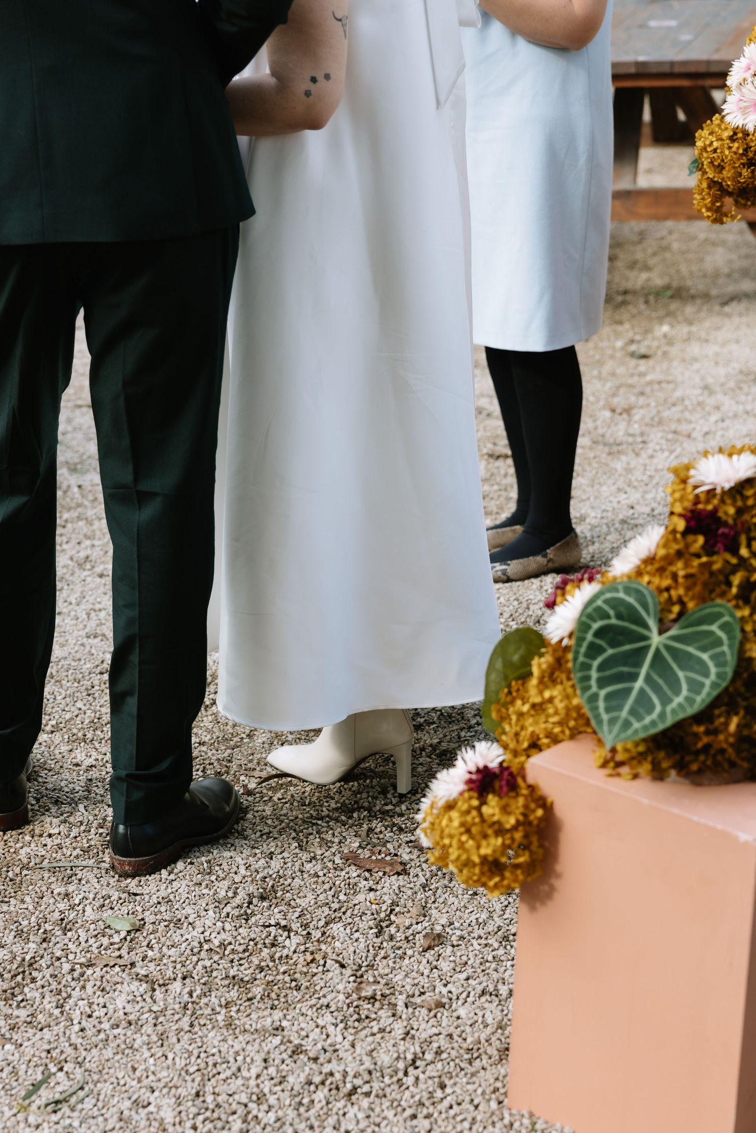 Wedding ceremony scene with bride in white dress and groom in dark suit standing near floral arrangements on gravel.