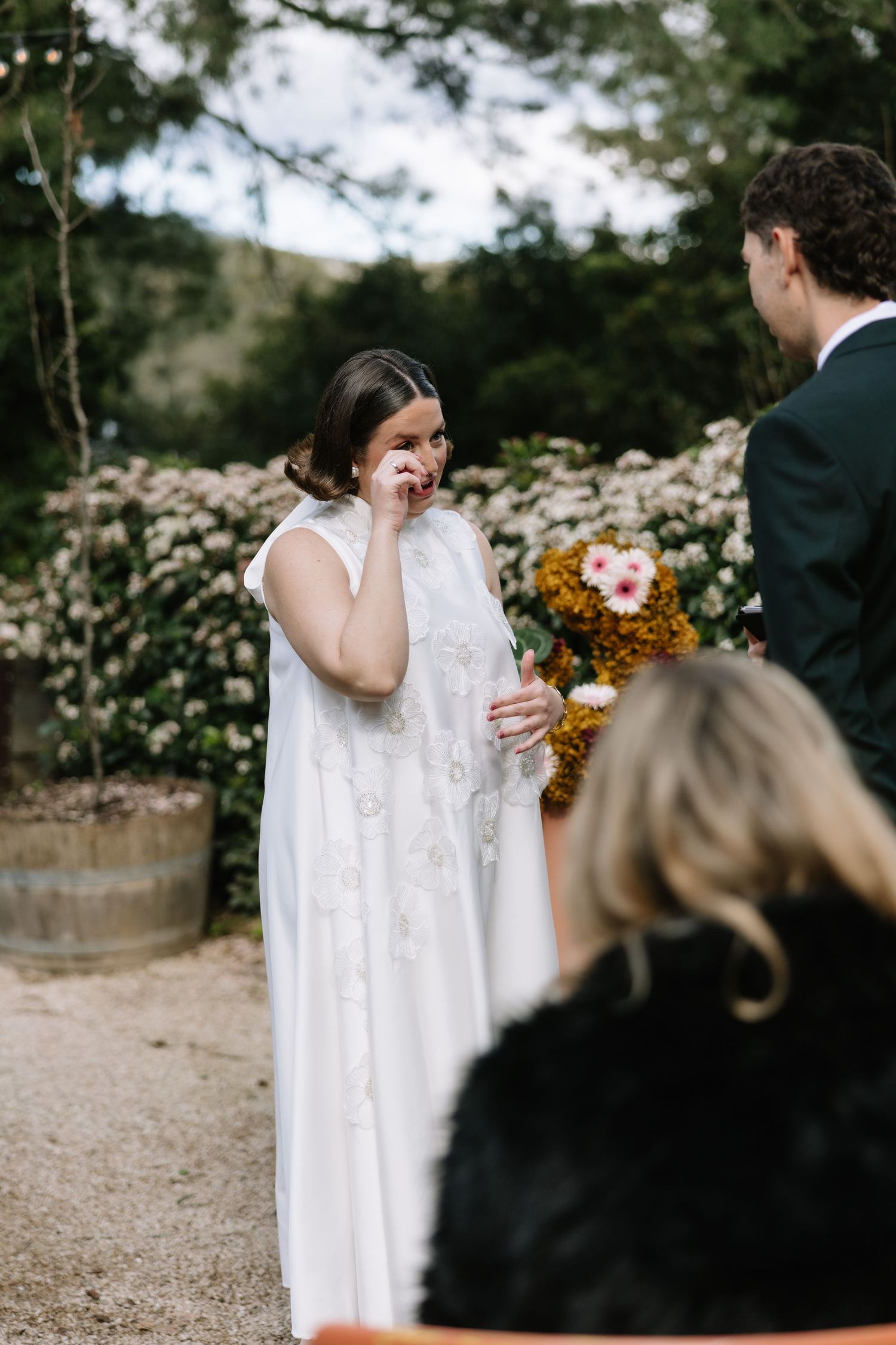 Emotional bride in white lace gown wiping tears during outdoor wedding ceremony surrounded by flowers.