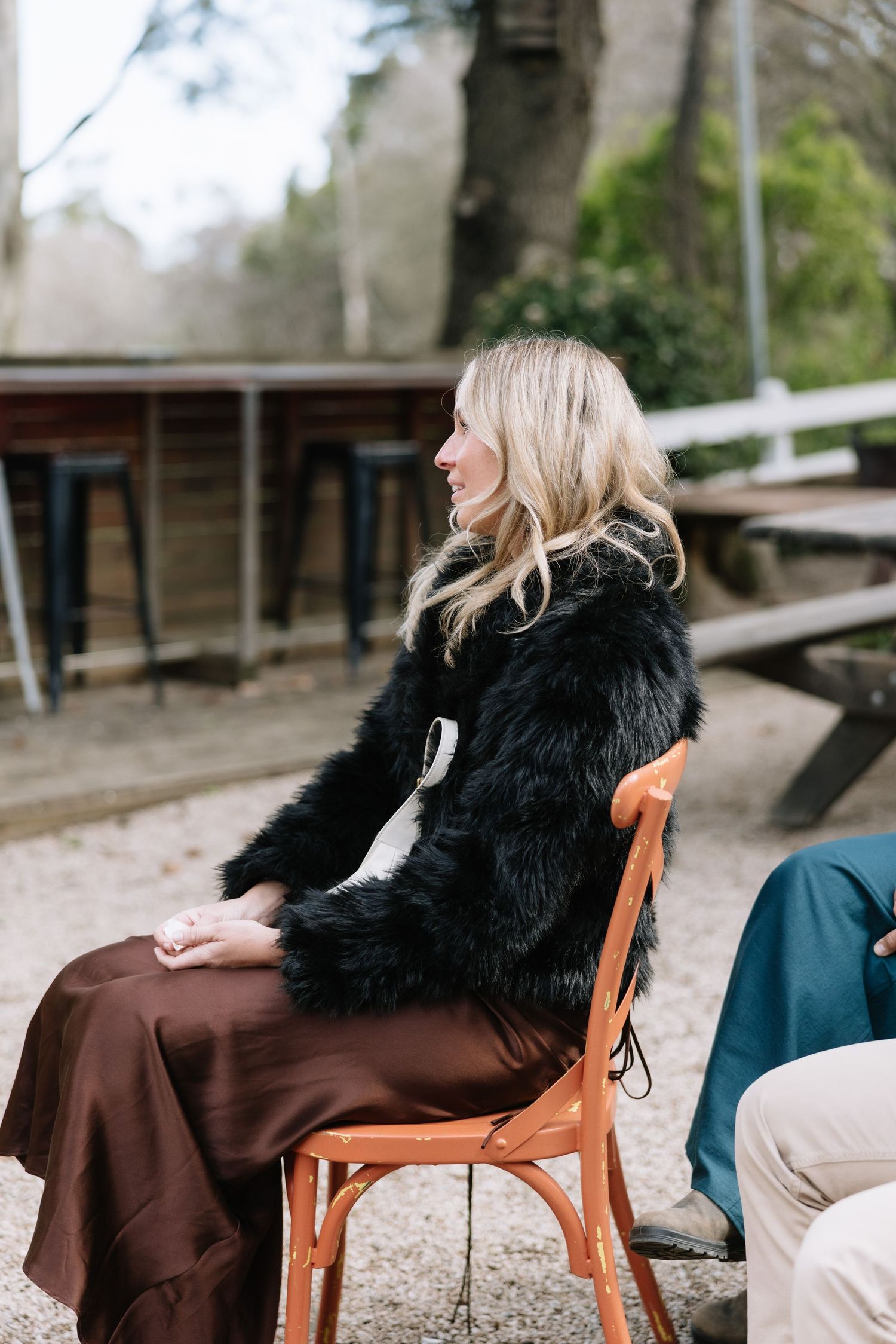 Blonde woman in black fur coat and brown satin skirt sitting on wooden chair outdoors at casual gathering.
