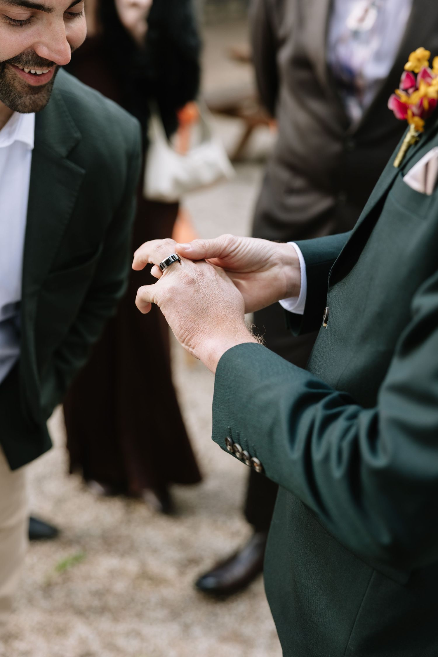 Two grooms in dark green suits exchange wedding rings during an outdoor ceremony.