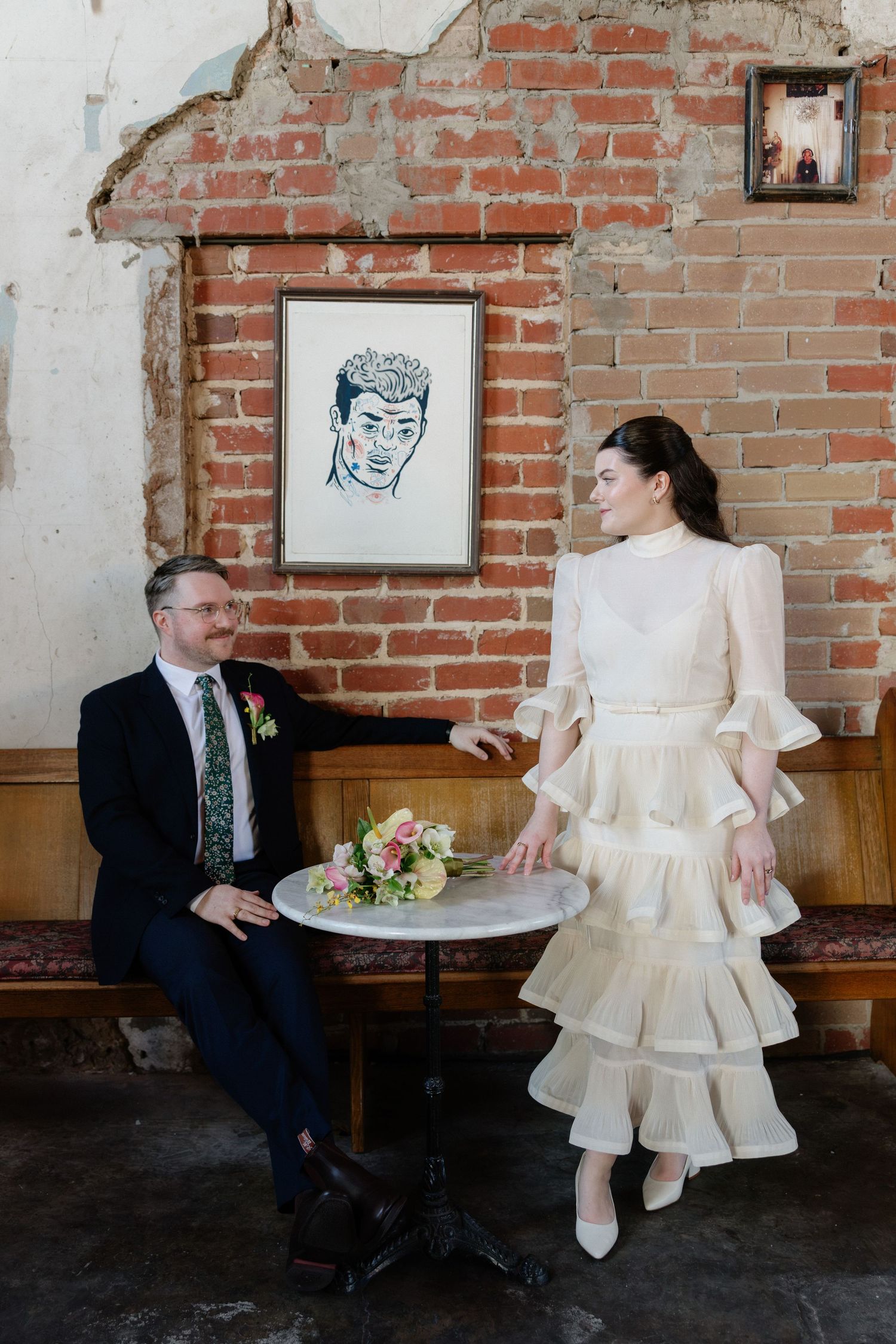 Bride and groom share a romantic moment at a marble table in front of an exposed brick wall with artwork.