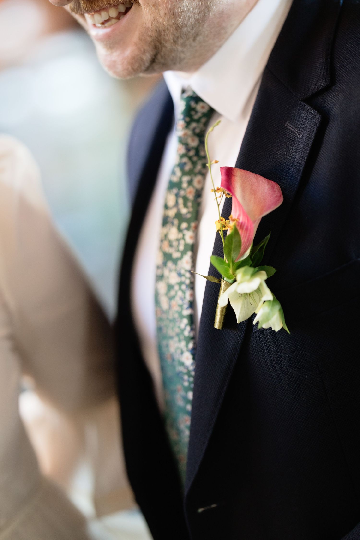 Close-up of groom's pink calla lily and white flower boutonniere on navy suit lapel with floral tie.