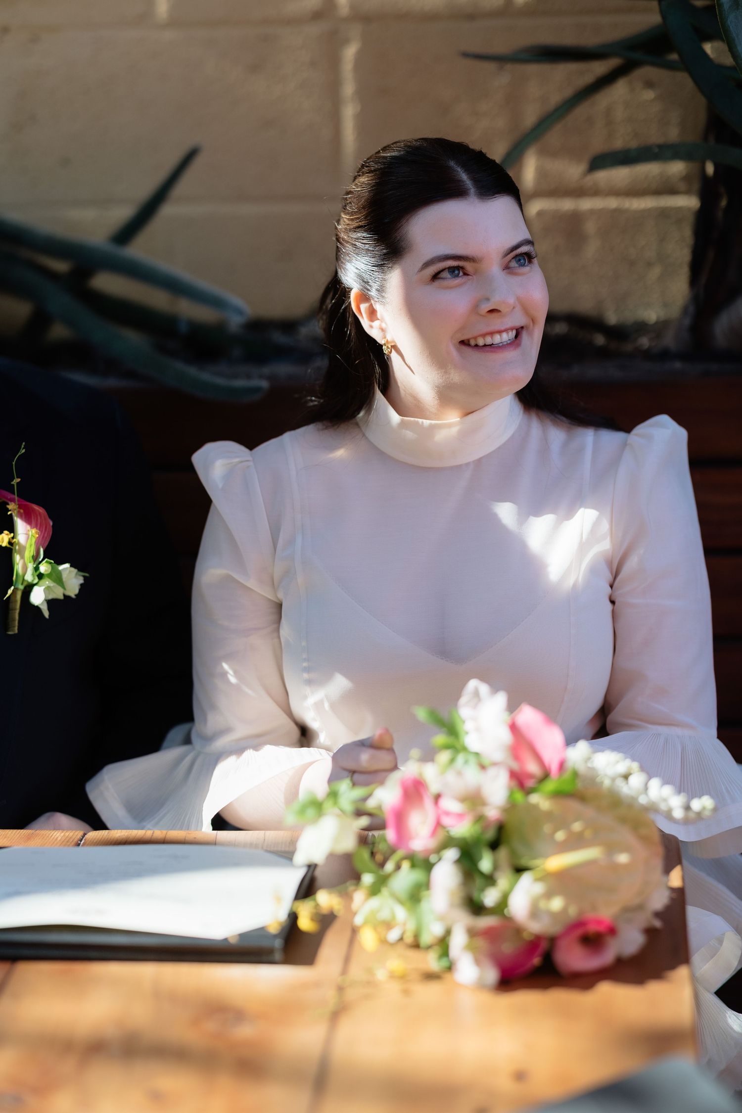 Smiling bride in white long-sleeve dress sitting at wooden table with pink floral bouquet during wedding ceremony.