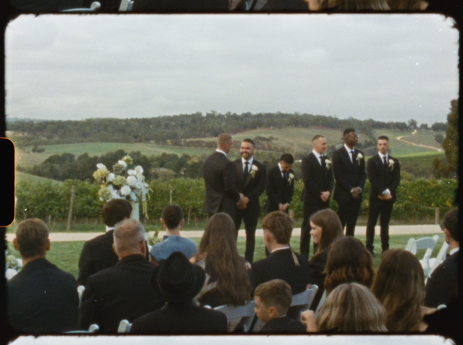 Wedding ceremony at a vineyard with groomsmen and guests gathered on a cloudy day.