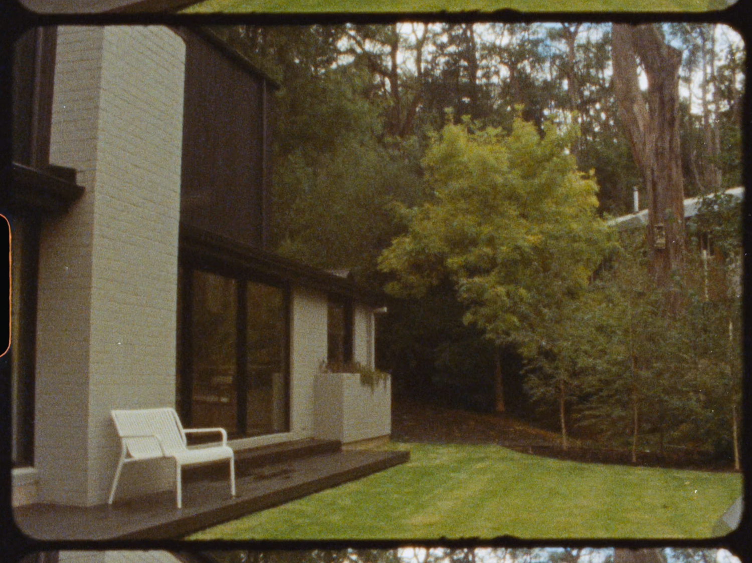 Modern home exterior with white chair on patio surrounded by lush green lawn and trees.
