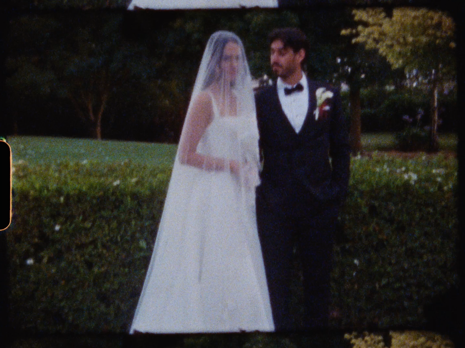 Bride in white wedding dress and veil standing with groom in black tuxedo in lush garden setting.