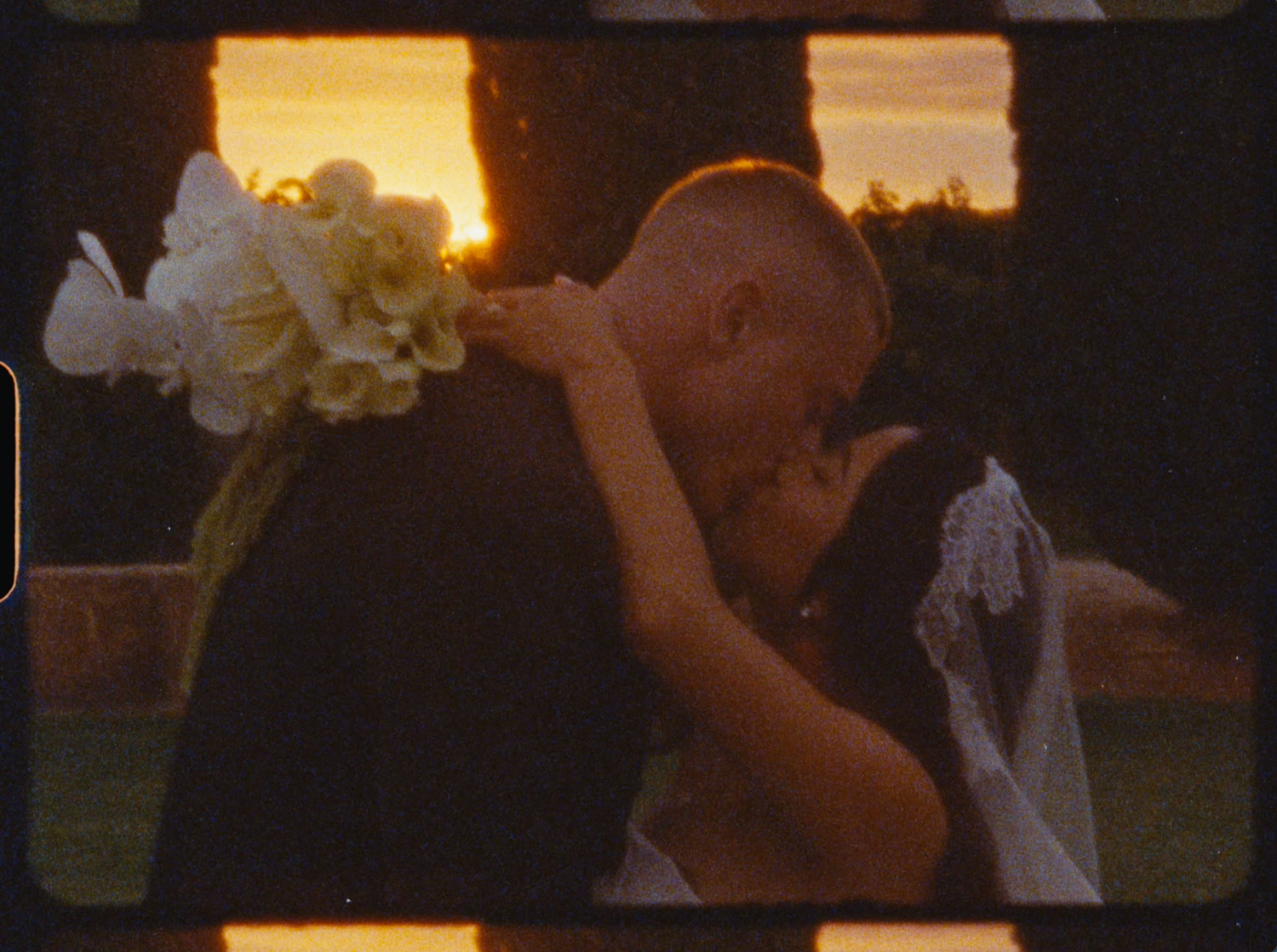 Bride and groom share a romantic sunset kiss surrounded by white flowers outdoors.