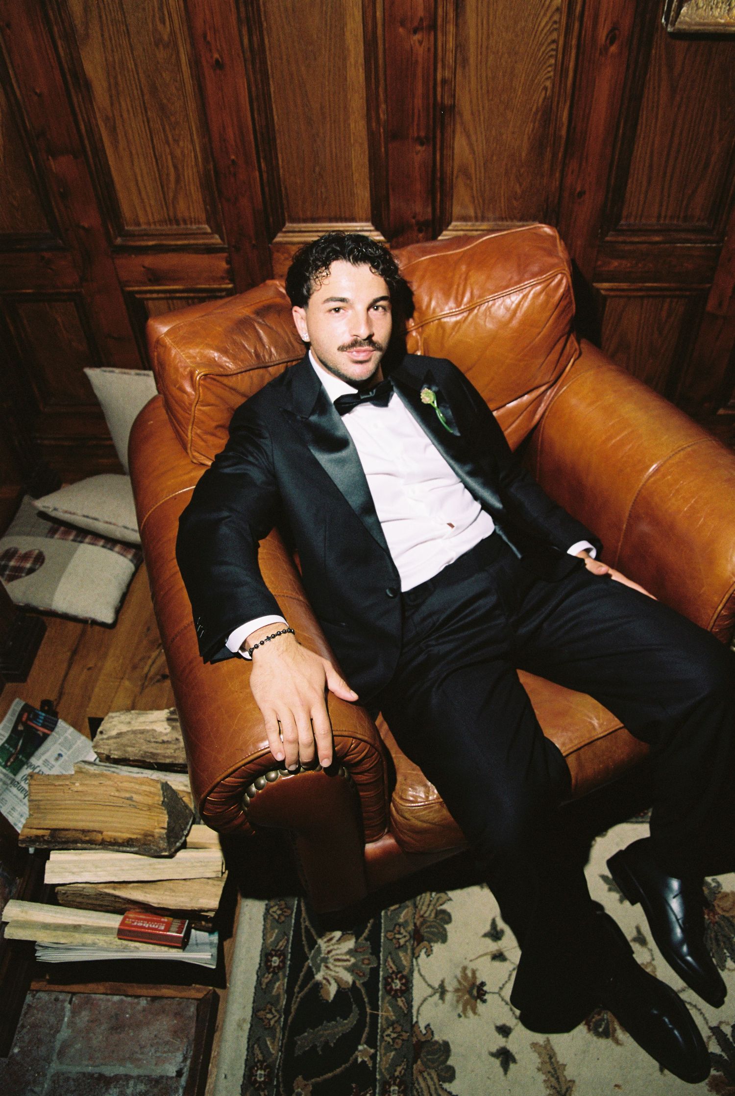 Groom in black tuxedo relaxing on a leather sofa surrounded by vintage books.