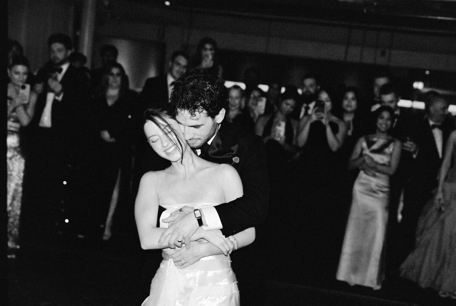 Bride and groom share their first dance surrounded by wedding guests in elegant black and white photo.
