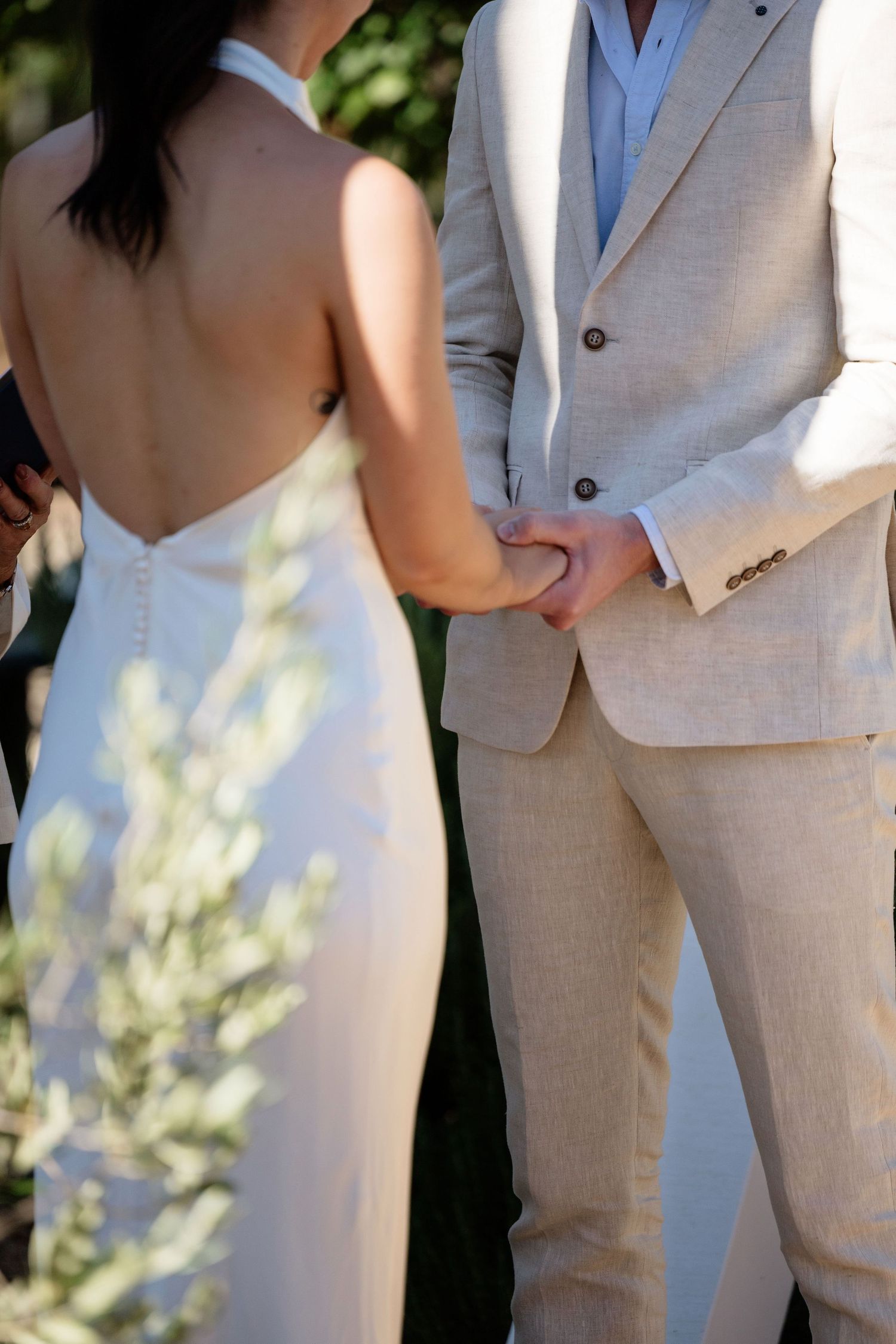 Bride in backless white halter gown holds hands with groom in beige linen suit during outdoor ceremony.