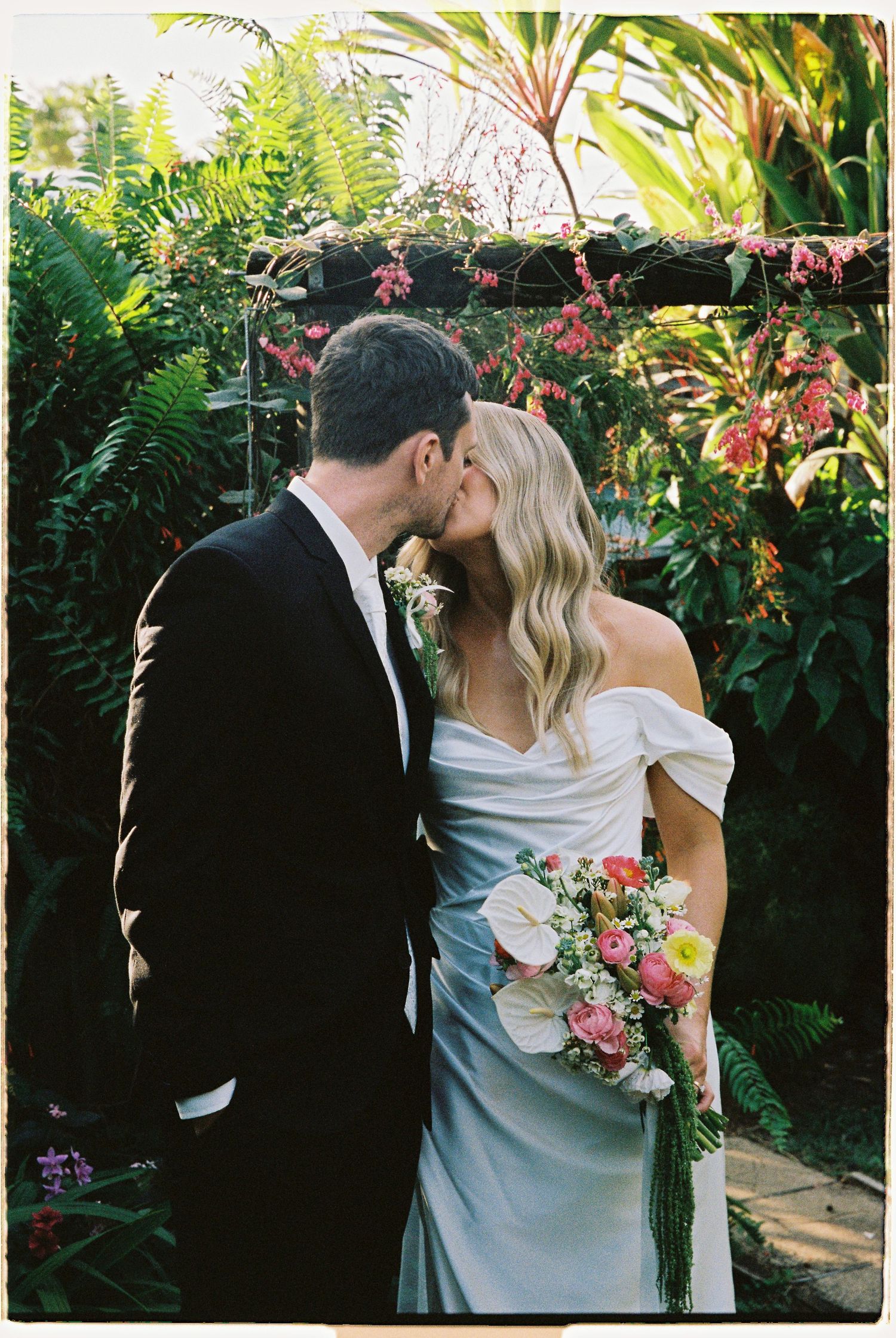 Bride and groom share a kiss surrounded by lush tropical greenery, bride holding a colorful bouquet in an off-shoulder gown.