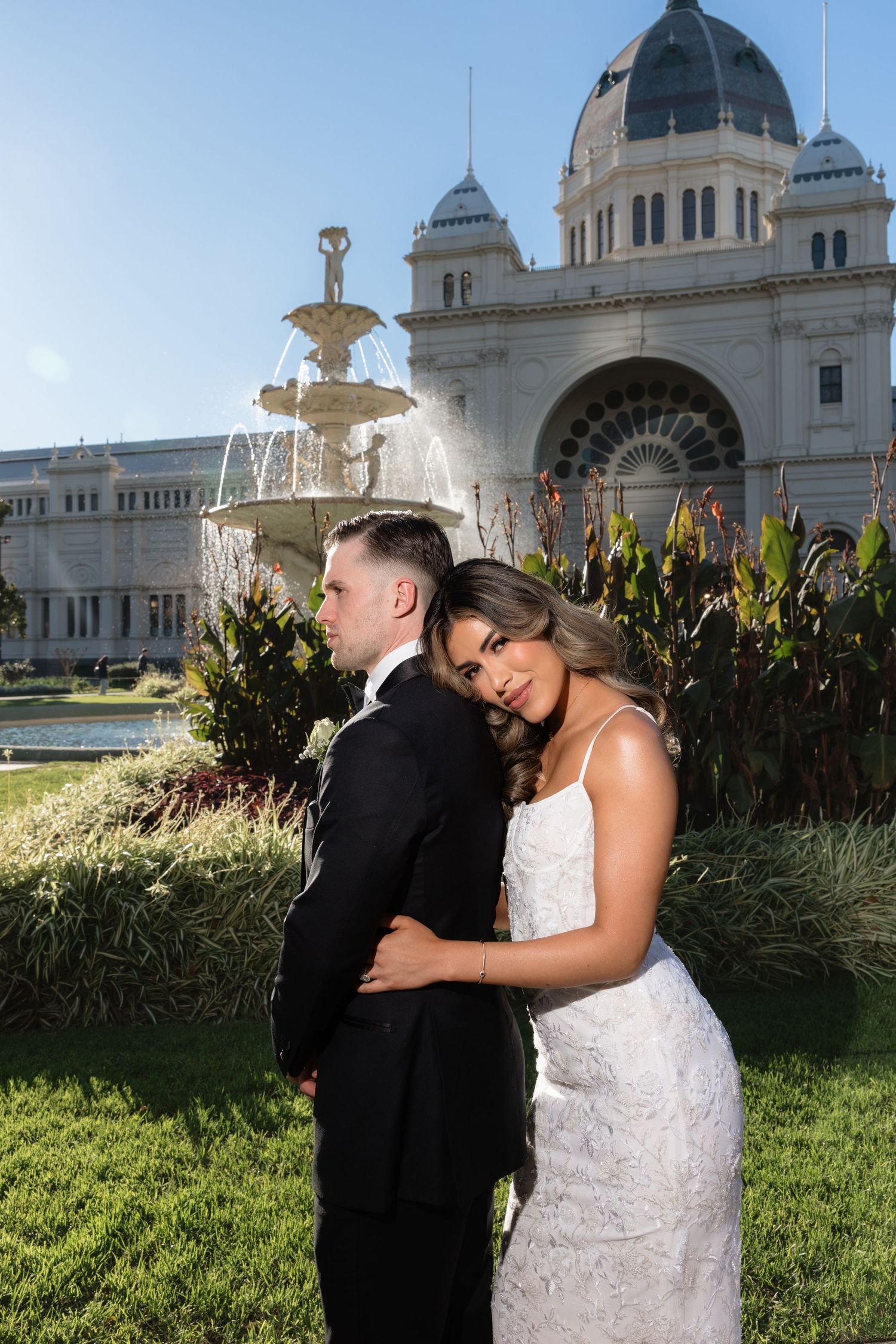 Bride leaning on groom's shoulder before grand Royal Exhibition Building fountain, bride in lace gown, groom in black suit.