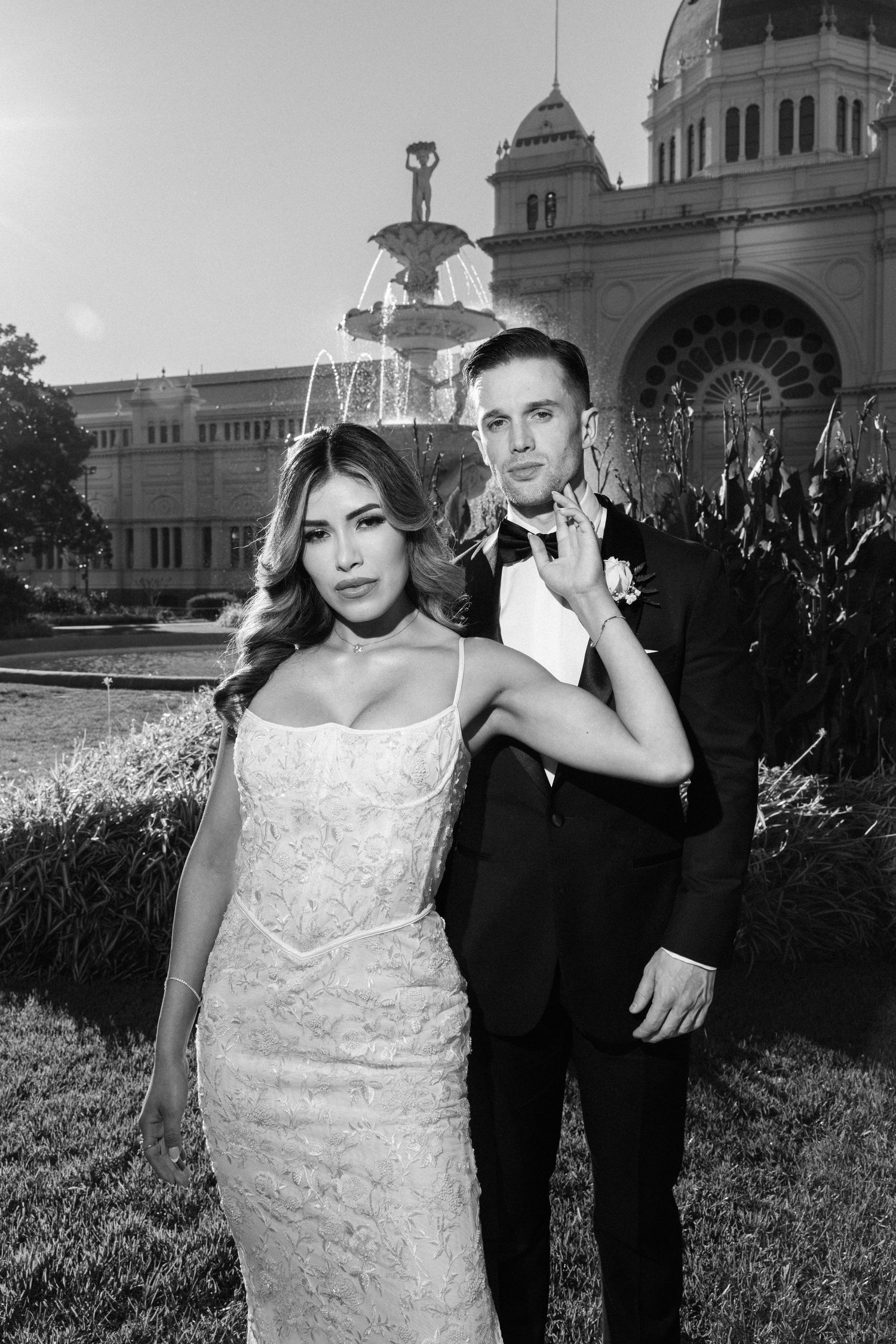 Black and white wedding photo of bride in lace gown and groom in tuxedo posing before ornate fountain and historic building.