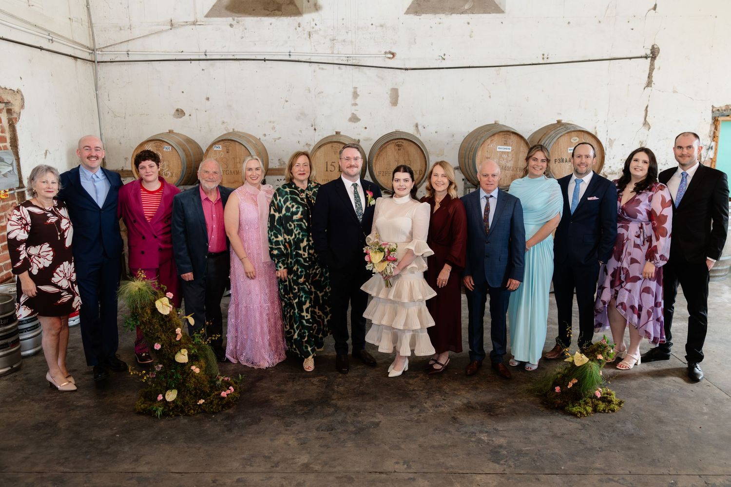 Wedding party group photo with bride in white dress posing with family in front of rustic wine barrels.