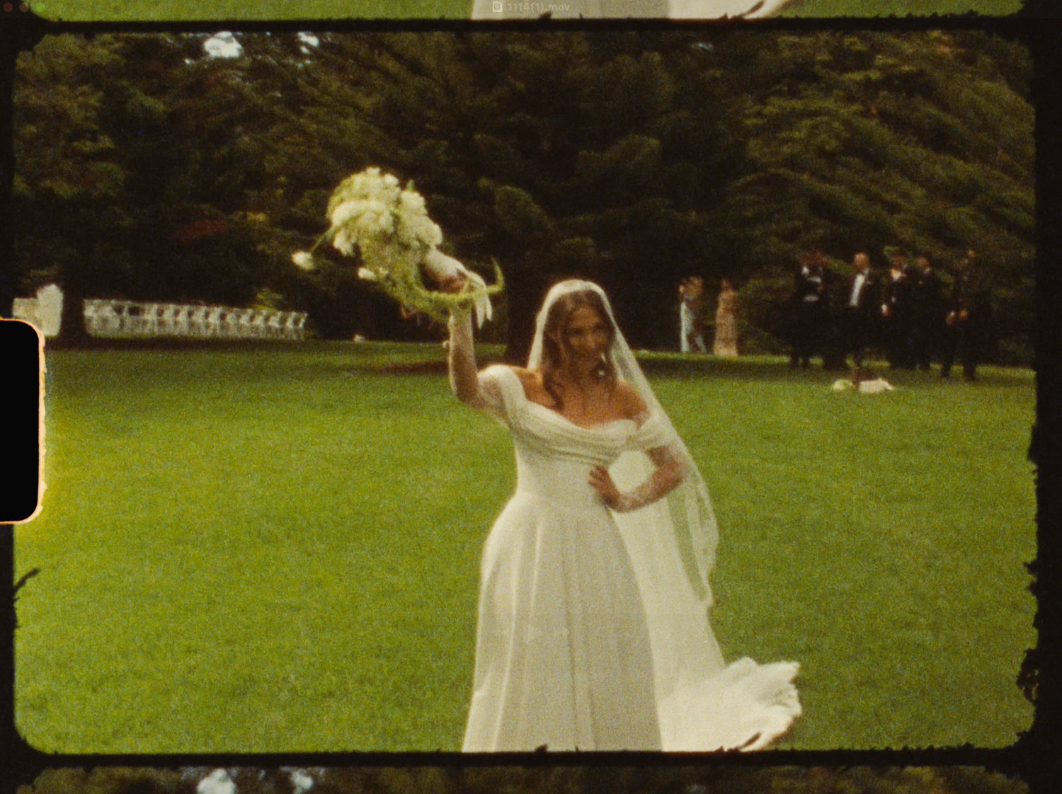 A bride in a white gown and veil raises her bouquet on a green lawn at an outdoor wedding ceremony.